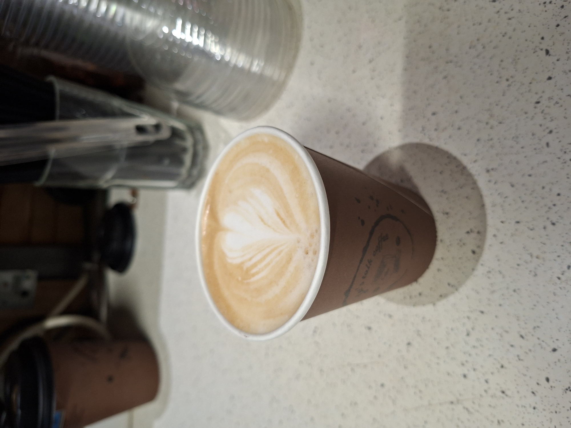 An overhead shot presents a latte in a brown paper cup, resting on a white, speckled countertop. The latte features a milk foam design resembling a heart or leaf. To the left of the cup, various cylindrical objects appear, possibly stacked or arranged. The background is a section of the speckled countertop. The location is Plovdiv, Bulgaria.
