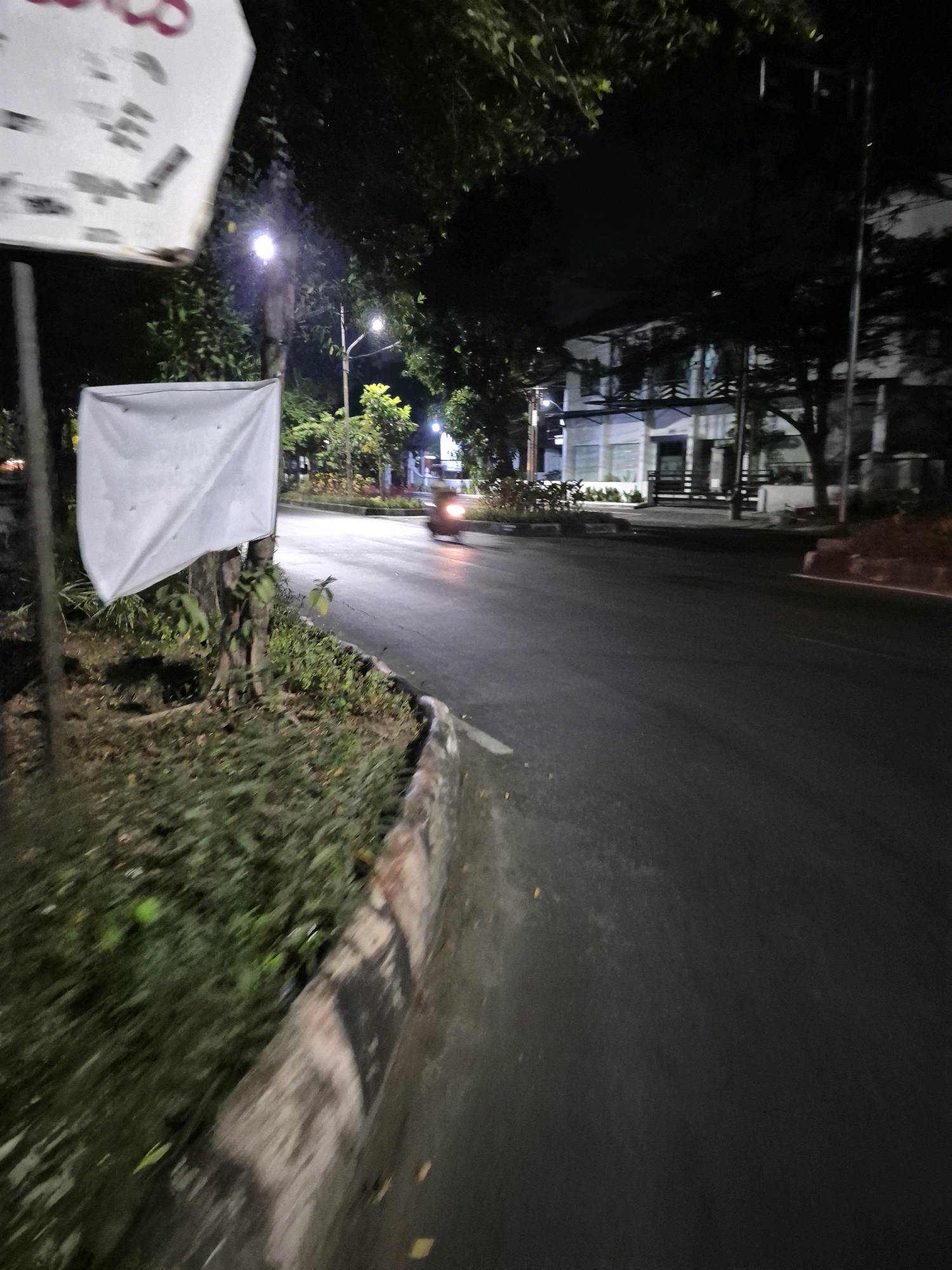A motorcycle is seen moving from left to right on a paved road at night. The road has a curb lined with vegetation. In the background, there is a building with multiple stories, illuminated by artificial lights. Streetlights are visible, casting a glow on the surroundings. A white banner is attached to a pole in the foreground, partially obscuring the view of a tree. The scene appears to be in a suburban or urban area of Surakarta, Indonesia, given the presence of tropical vegetation and the architectural style of the building. The image captures a typical nighttime street scene with ambient lighting from streetlights and the building.