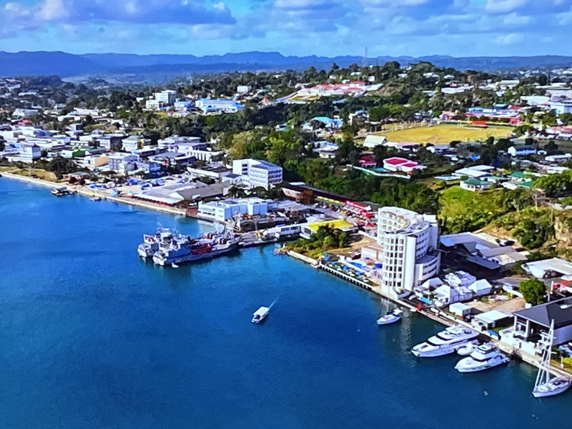 This is an aerial drone shot of a coastal city with a harbor. The water is a deep blue, and a large ship, several smaller boats, and a white speedboat are visible in the water. The city is built along the coast and extends inland up a hill. Buildings are a mix of white, red, and gray structures, with some appearing to be commercial or residential. Green trees and vegetation are scattered throughout the city. In the background, mountains are visible. The sky is blue with some white clouds. The scene appears to be a sunny day. The location is not Kharkiv, Ukraine, as Kharkiv is an inland city.