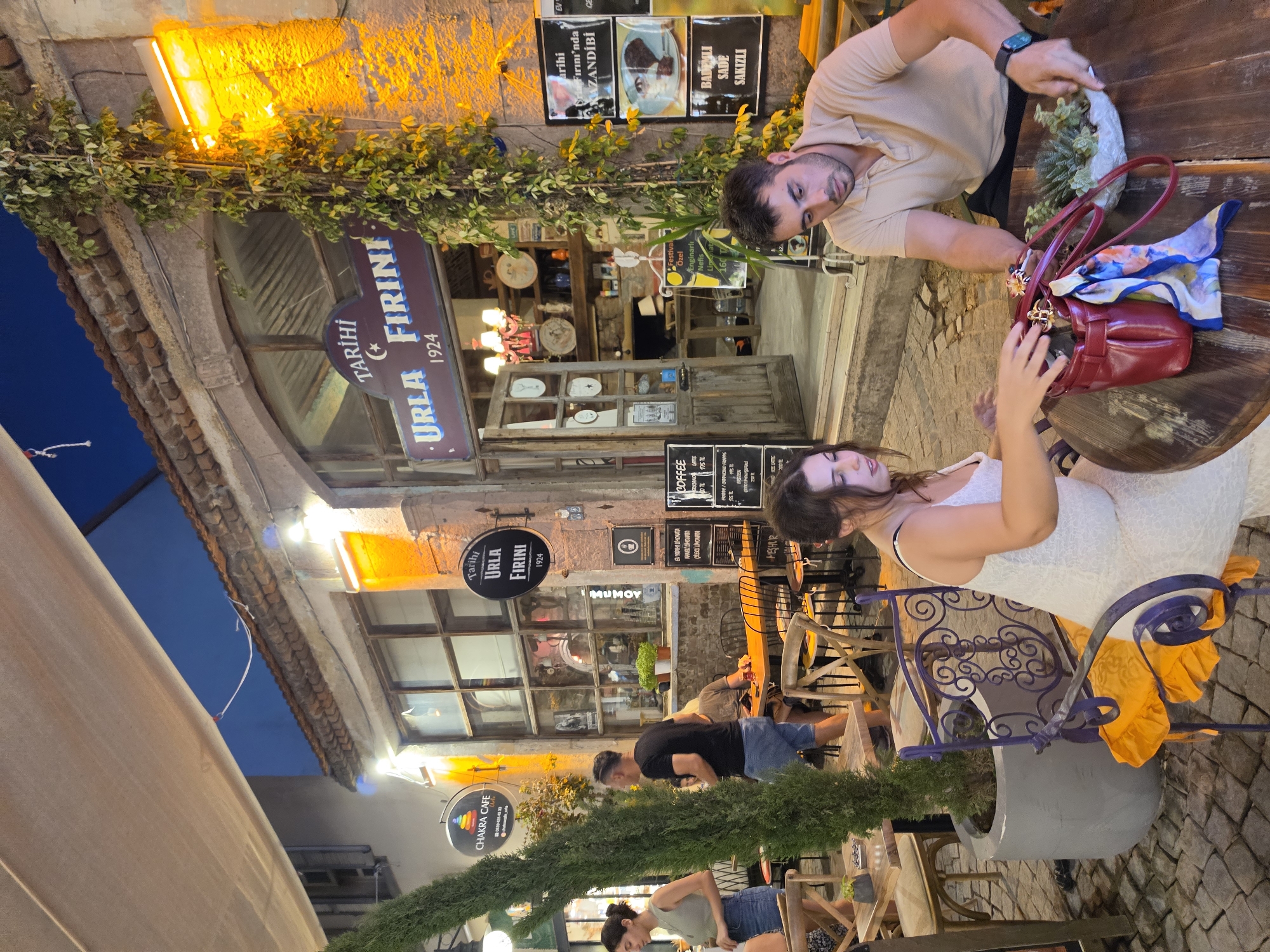 The image depicts an outdoor seating area of a cafe or restaurant in Urla, Turkey. The establishment features a sign that reads "Tarihi Urla Fırını 1924," indicating it is a historic bakery. In the foreground, a man and a woman are seated at a round wooden table. The man, wearing a tan polo shirt and black shorts, is looking down at the table. The woman, wearing a white lace dress, is reaching into a red handbag. A small succulent plant in a white pot is also on the table, along with a colorful scarf.
In the background, more tables and chairs are visible. The building facade is made of stone and has large windows. Green ivy is growing up the side of the building. Several other people are seen walking and sitting in the area. A tall, conical tree stands to the left of the scene. The lighting suggests it is evening, with warm artificial lights illuminating the area. The ground is paved with cobblestones. Signs in Turkish are visible on the building, advertising items like "coffee" and various food items. The overall atmosphere appears to be lively and social.