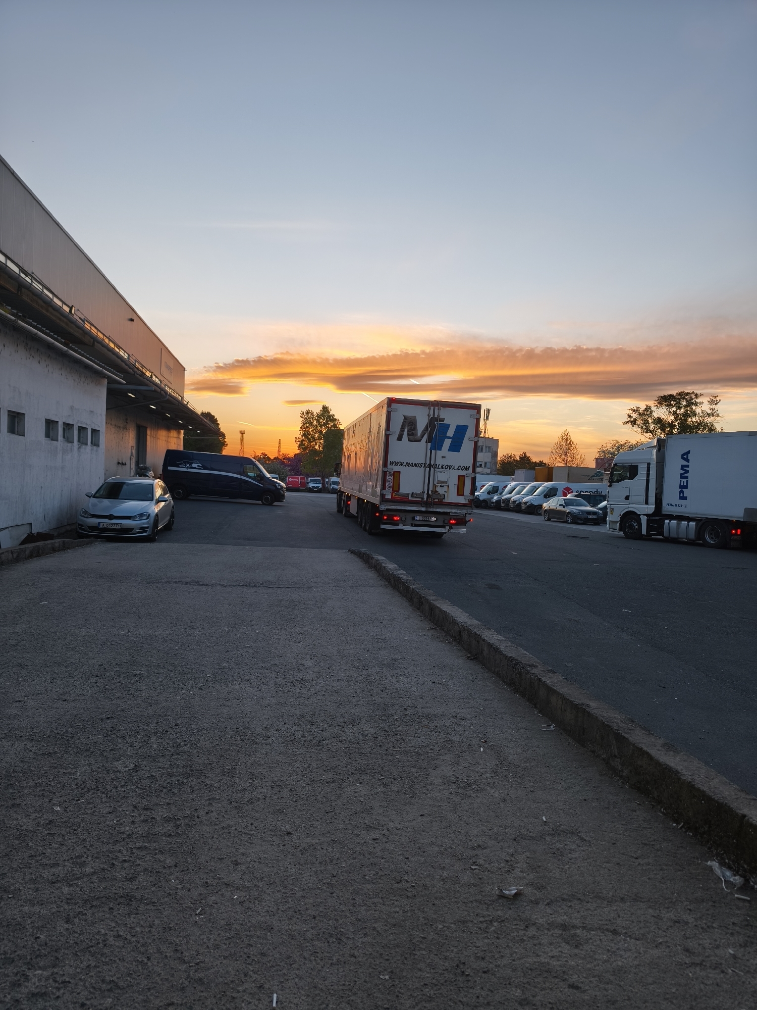 The image depicts an industrial or logistics area in Burgas, Bulgaria, during either sunrise or sunset, characterized by a vibrant sky transitioning from warm orange and yellow clouds at the horizon to a clear light blue overhead.
The primary subjects include several commercial vehicles and a large warehouse-type building. On the left, a long, white and grey industrial building features loading docks. A silver Volkswagen Golf with Bulgarian license plate "A 0127 PH" is parked adjacent to this building, followed by a dark van with graphics on its side.
In the center and right, an asphalt road and parking area hold several semi-trailer trucks and vans. A white semi-trailer truck, displaying the "MH" logo and "WWW.MANISTHALKOVA.COM" on its rear, is parked facing away from the viewer, showing a Bulgarian "C" series license plate. To its right, another white semi-trailer truck is visible with "PEMA" written on its side. In the background, multiple white delivery vans, some identifiable with the "Speedy" logo, are also parked.
The scene is quiet, with no people visibly engaged in activity, suggesting a time outside of peak operational hours. The ground is covered in asphalt, separated by a concrete curb. Distant trees and other buildings are visible against the colorful sky. The striking natural light of the golden hour bathes the scene, creating warm tones and soft shadows.