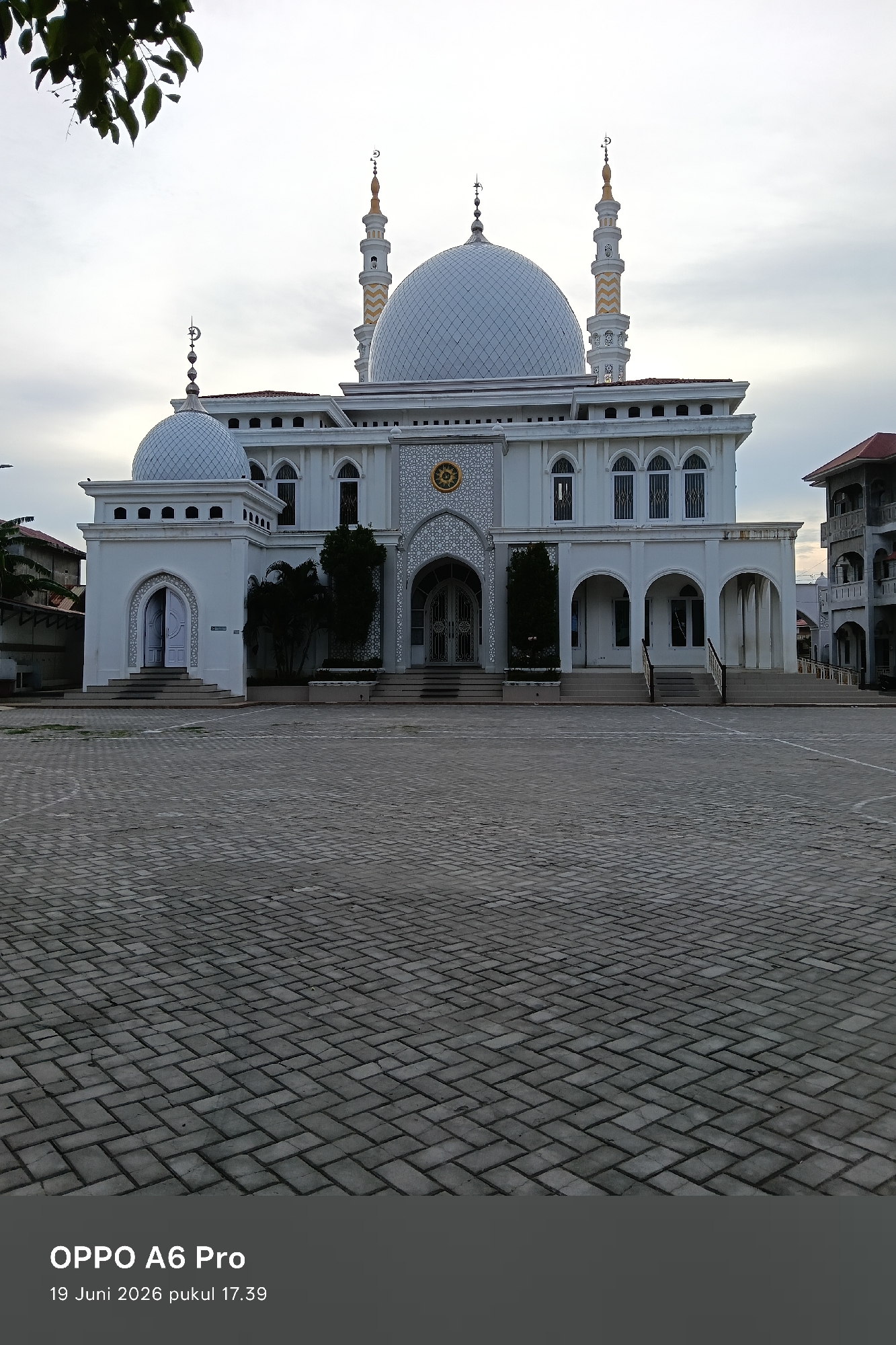 The image shows the exterior of the Islamic Center of Lhokseumawe, Indonesia. The building features a large white dome and two white minarets with gold accents. The facade is white with arched windows and decorative elements. In front of the building is a paved courtyard with a herringbone pattern. The sky is overcast, and the lighting suggests it is late afternoon or early evening. There are no people visible in the image. The text at the bottom of the image indicates it was taken with an OPPO A6 Pro on June 19, 2026, at 17:39.