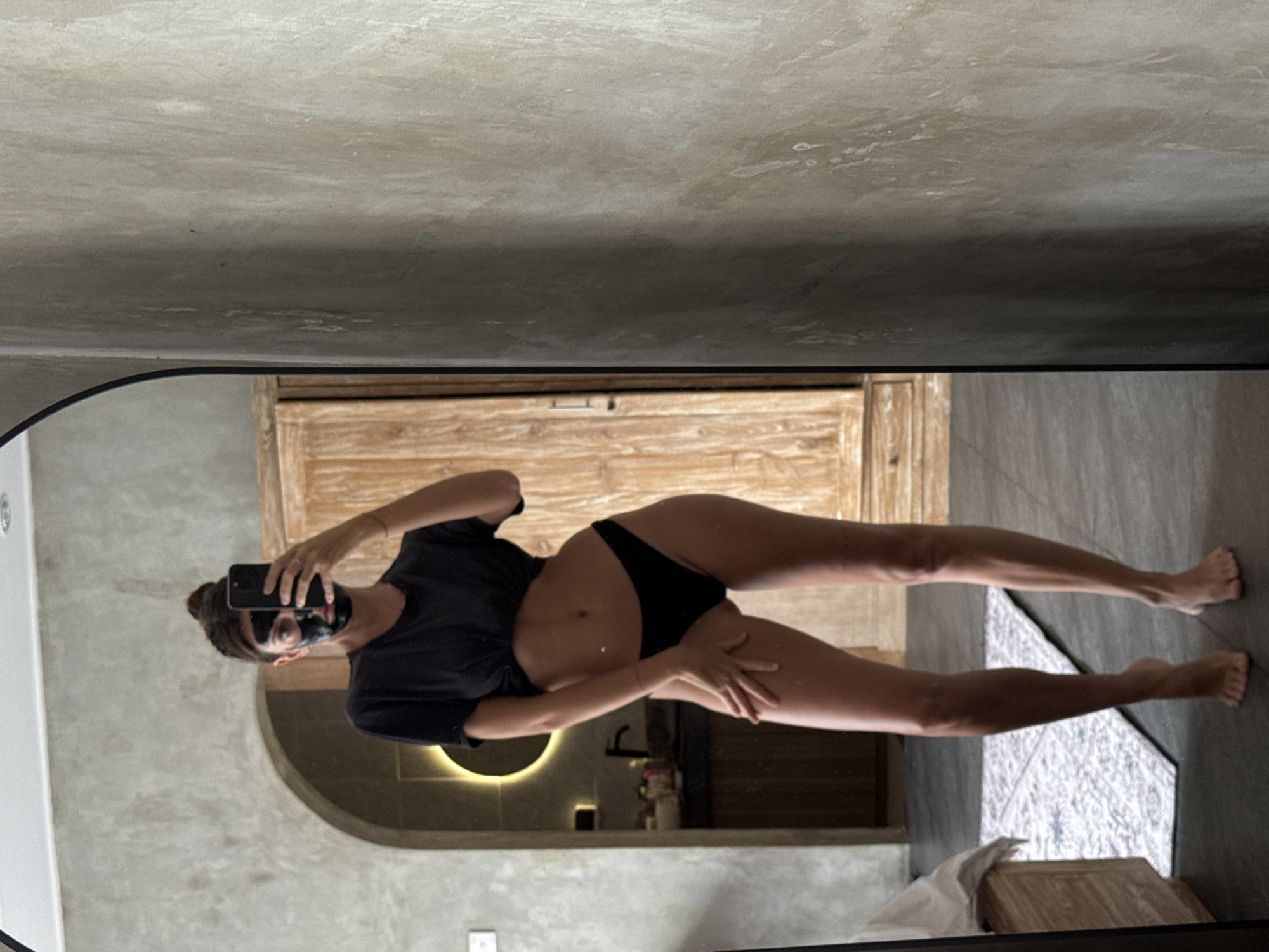 A person with dark hair pulled into a bun is standing in front of a mirror, taking a selfie. They are wearing a black crop top and black underwear, and a black face mask. The background shows a bathroom with a sink and a wooden cabinet. The scene appears to be in a modern home or hotel in North Kuta, Indonesia.