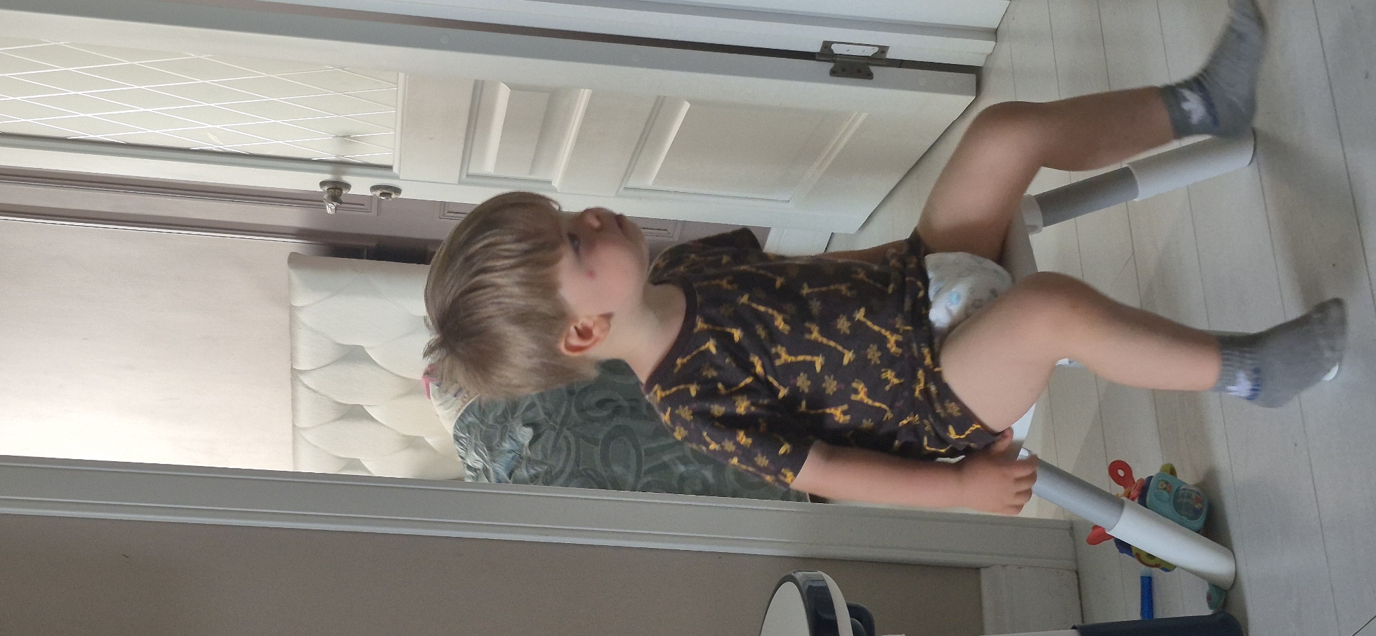 A young child with blonde hair is sitting on a white and grey baby walker. The child is wearing a dark t-shirt with a giraffe pattern and grey socks. The child is facing away from the camera, looking up. A diaper is visible under the t-shirt. The background shows a white door with diamond-shaped patterns, a white quilted mattress, and a patterned rug. The floor is light-colored wood. A colorful toy is visible near the walker. The lighting suggests it is daytime.