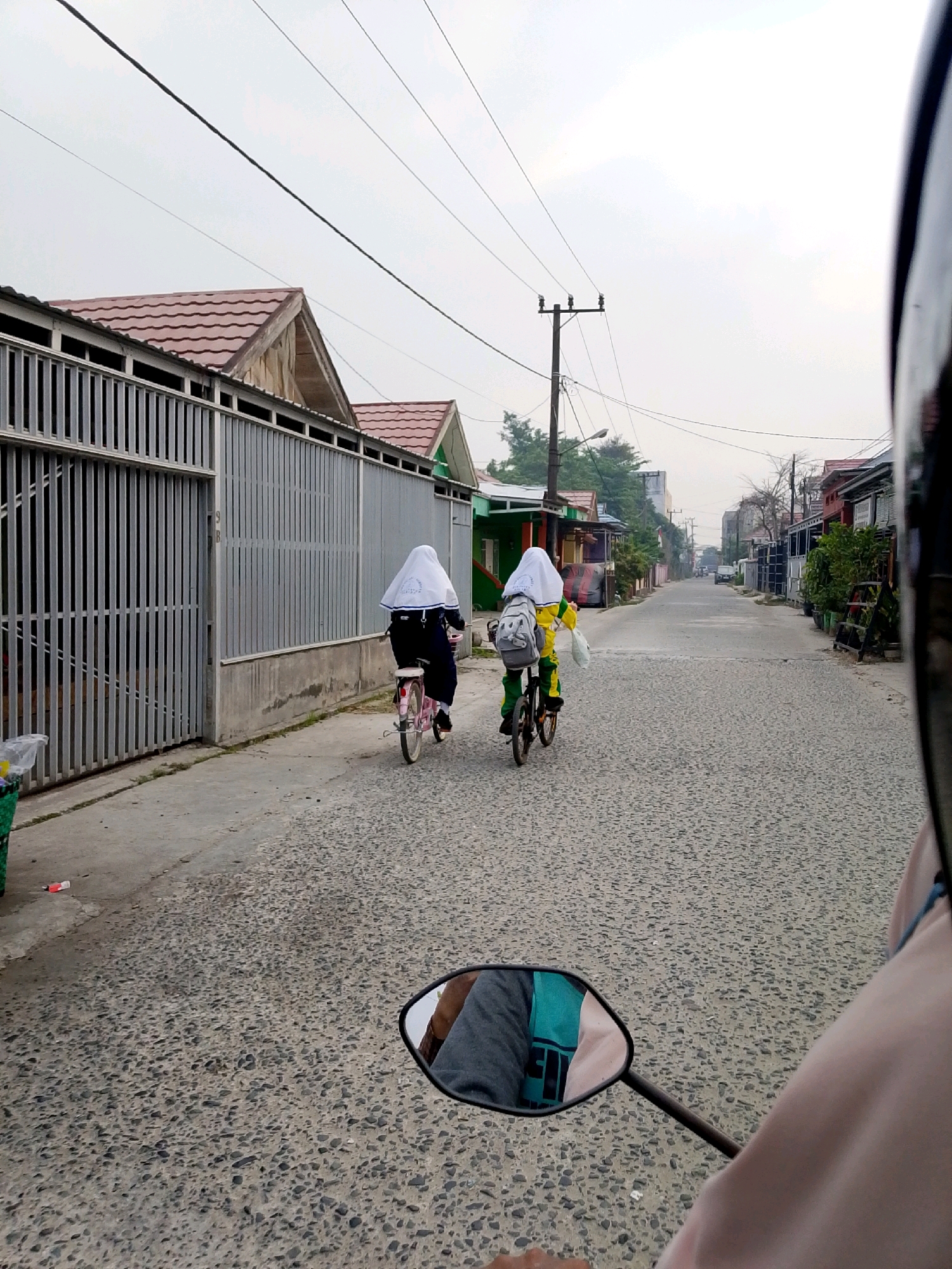 In Gambut, Indonesia, two young individuals, presumably students, are riding bicycles down a paved street lined with houses and fences. The person on the left is wearing a white hijab and dark clothing, while the person on the right is in a yellow hijab and matching outfit. Both have backpacks. The street is made of asphalt with visible pebbles. Power lines stretch across the overcast sky above. In the foreground, the reflection in a motorcycle's rearview mirror shows the shoulder of a person wearing a pink jacket and a glimpse of their grey sleeve and a turquoise bag. The environment suggests a residential area.