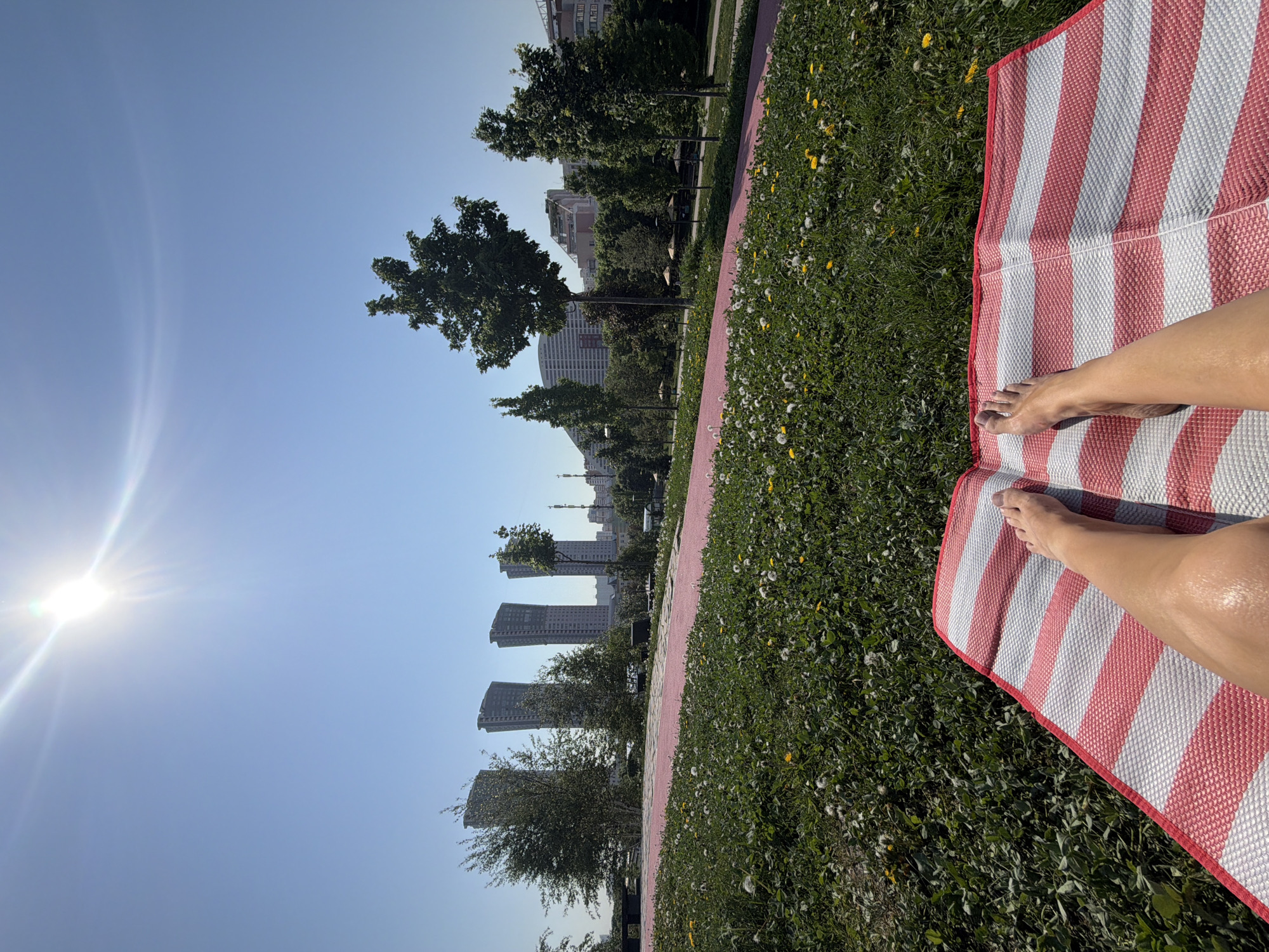A sunny day in Khimki, Russia, shows a person lying on a red and white striped mat on a green grassy area. The person's legs and feet are visible on the mat. In the background, there is a path and several high-rise buildings are visible through the trees. The sun is shining brightly in the sky.