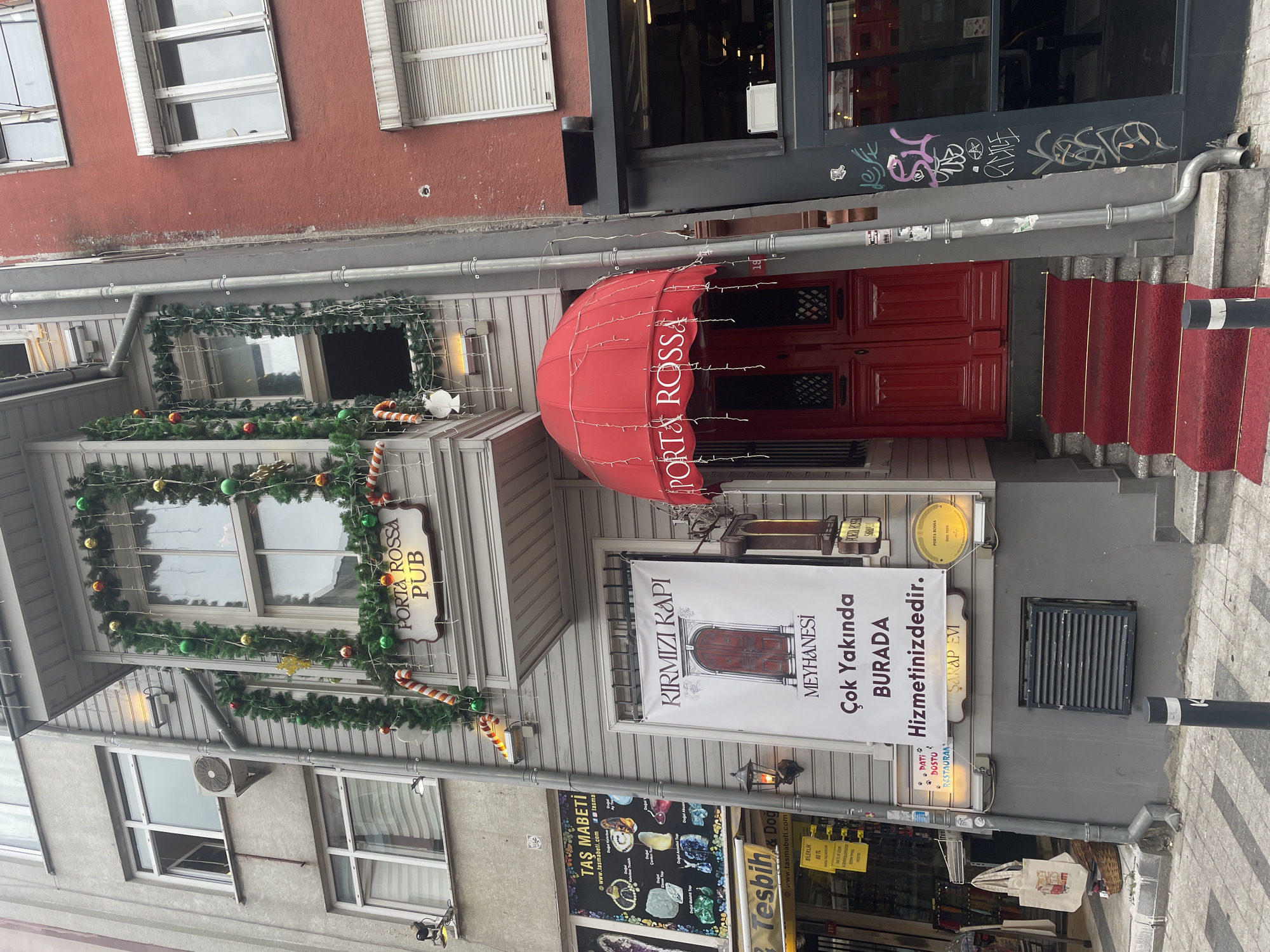 The image shows a narrow street in Kadıköy, Turkey, with a building facade decorated for Christmas. A sign reads "Porta Rossa Pub" above a window. Below, a large banner announces "Kirmizi Kapi Meyhanesi Cok Yakinda Burada Hizmetinizdedir" which translates to "Red Door Tavern Coming Soon Here We Serve You." A red awning covers the entrance to "Porta Rossa" with a red door. A red carpet leads up to the entrance. To the left of the building, a shop displays jewelry and gemstones. The building's exterior is grey wood siding, with a red-bricked building to the right. Graffiti is visible on the wall to the right of the red door.