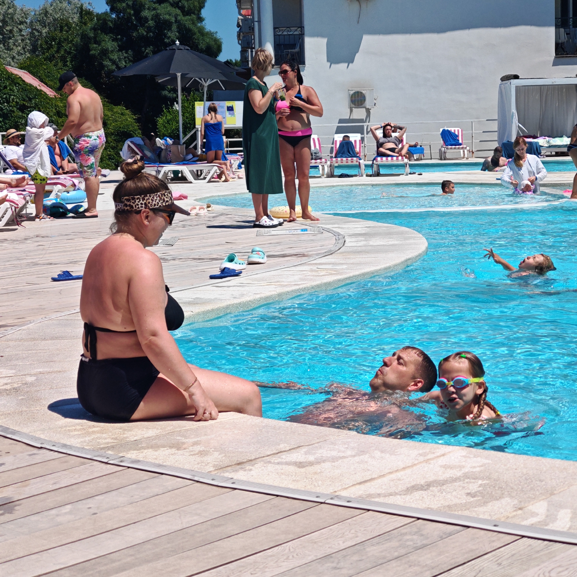 A group of people are relaxing by a swimming pool on a sunny day. In the foreground, a woman in a black bikini sits on the edge of the pool, facing away from the camera. She wears a leopard print headband and sunglasses. Behind her, a man and a young girl in swimsuits are in the water. The man is lying on his back, looking up, while the girl wears goggles and appears to be playing in the water.
In the middle ground, two women are standing near the pool. One is wearing a green dress and the other a pink and black bikini. Several other people are visible lounging on chairs and in the pool. The setting appears to be a resort or hotel, with white buildings and umbrellas visible in the background. The overall atmosphere is relaxed and summery. There is no visible text in the image.
