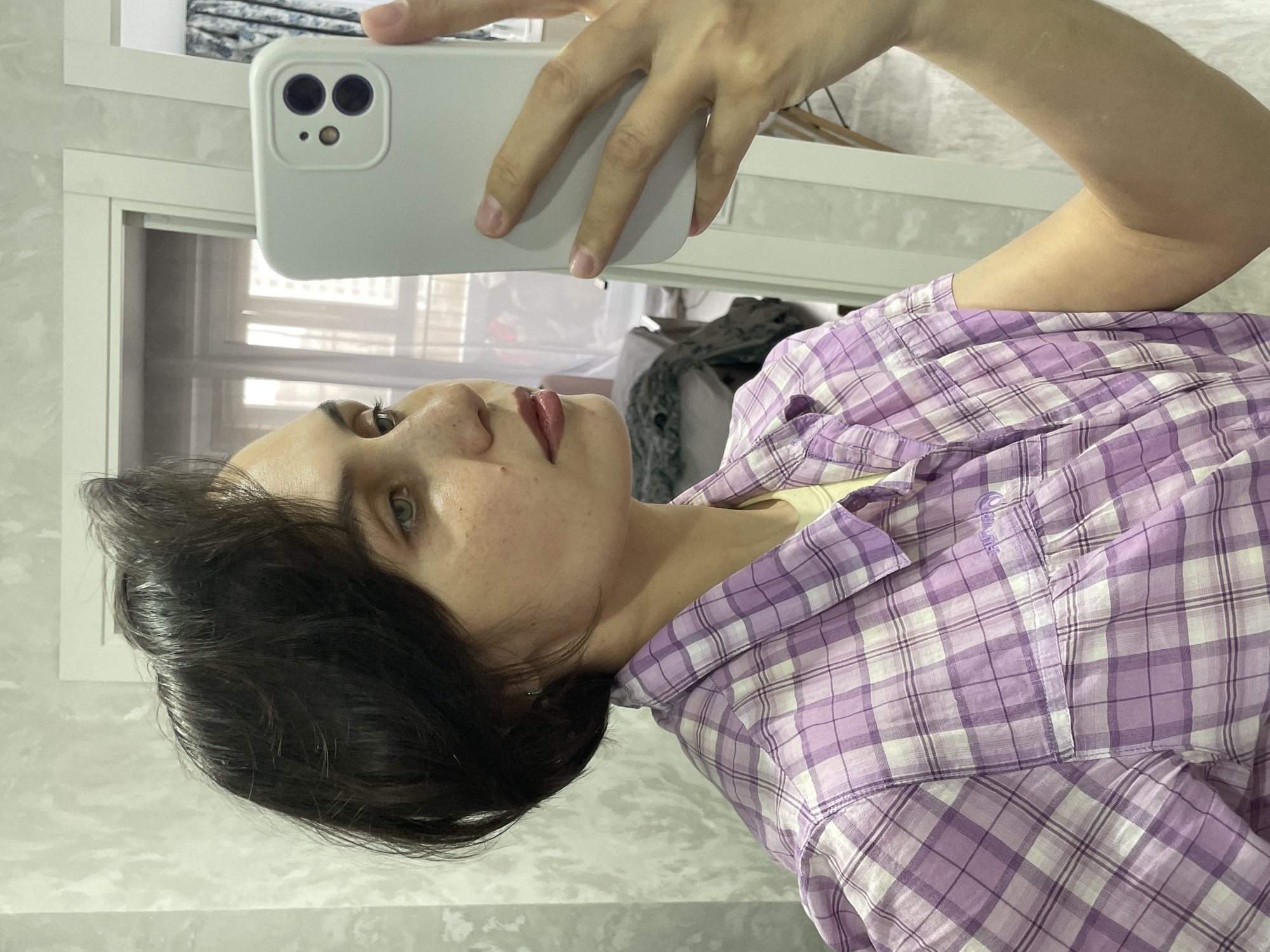 A selfie is captured in a mirror, showing a person with dark hair and freckles, wearing a purple and white plaid shirt. The individual holds a white-cased smartphone with visible dual camera lenses, aiming it towards their reflection. The person has dark or red lipstick and appears to have light-colored eyes. A subtle yellow garment is visible underneath the plaid shirt.
The background, seen in the mirror's reflection, suggests an indoor setting with light-colored, possibly patterned walls. A window or door frame with blinds is reflected in the upper left corner. Another reflection in the mirror shows a rack or hanger with some clothing. The overall lighting indicates it is likely daytime. The photo is oriented ninety degrees clockwise, showing the subject on their side. Faint text reading "Outdoors" is visible on the plaid shirt near the collar. The scene takes place in Izhevsk, Russia.