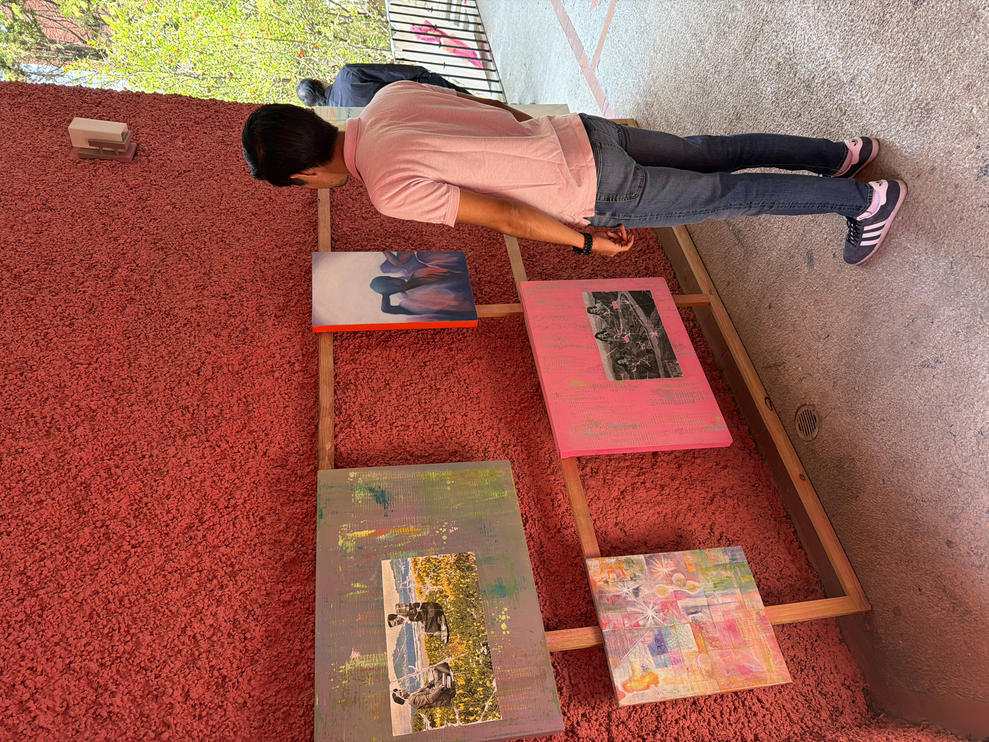 A person wearing a light pink shirt, blue jeans, and Adidas sneakers is standing and looking down at four paintings in a wooden frame, which is placed on a red textured ground. Another person is standing in the background. The scene is located in San Andrés Cholula, Mexico, as indicated by the prompt. The wall behind the paintings is also red textured.