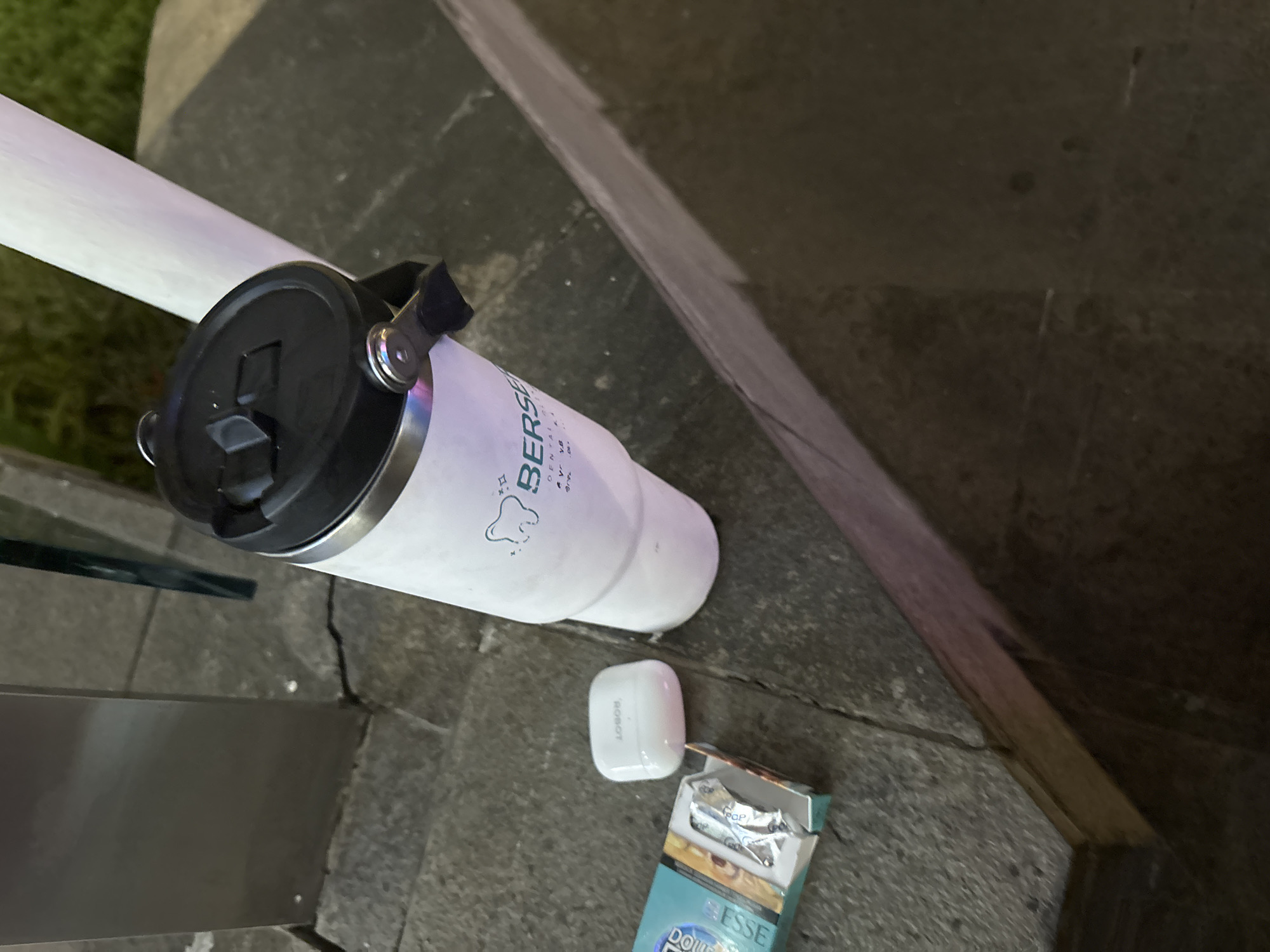 The image shows a close-up, eye-level view of several objects resting on a concrete surface, possibly an outdoor seating area or pavement in North Jakarta, Indonesia. In the foreground and to the left, a partially opened pack of "ESSE" cigarettes is visible. Inside the pack, the foil wrapper of cigarettes can be seen. Next to the cigarette pack is a small, white, oval-shaped object, which appears to be a wireless earbud charging case with "ROBOT" printed on it. To the right of these items is a tall, white insulated tumbler with a black lid. The tumbler has a logo featuring a stylized tooth and the word "BERS...". The background is blurred, showing a gray metal structure on the left and a white cylindrical object, possibly a pole or support, on the right. The ground surface is textured concrete with visible cracks and dark shadows, suggesting it might be evening or the area is dimly lit. The overall scene depicts a collection of personal items left unattended.