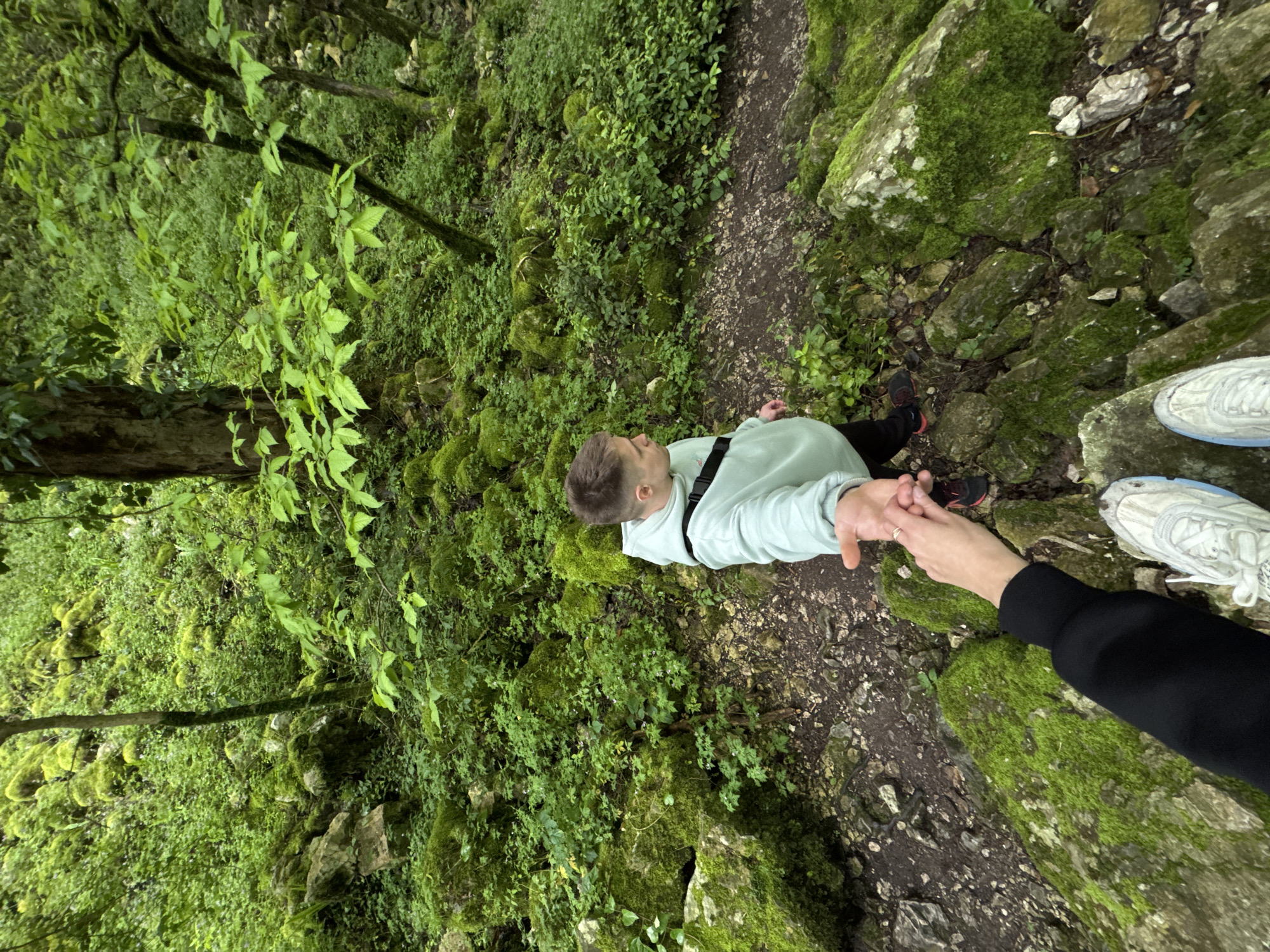 A high-angle shot captures a man standing on a dirt path in a green forest. The man is wearing a light green sweatshirt and black pants with black shoes. He is holding hands with someone whose arm and part of their legs and white sneakers are visible on the right. The environment is lush with green moss covering rocks and trees. The dirt path meanders through the scene. This scene occurs in Tsabal, Abkhazia.