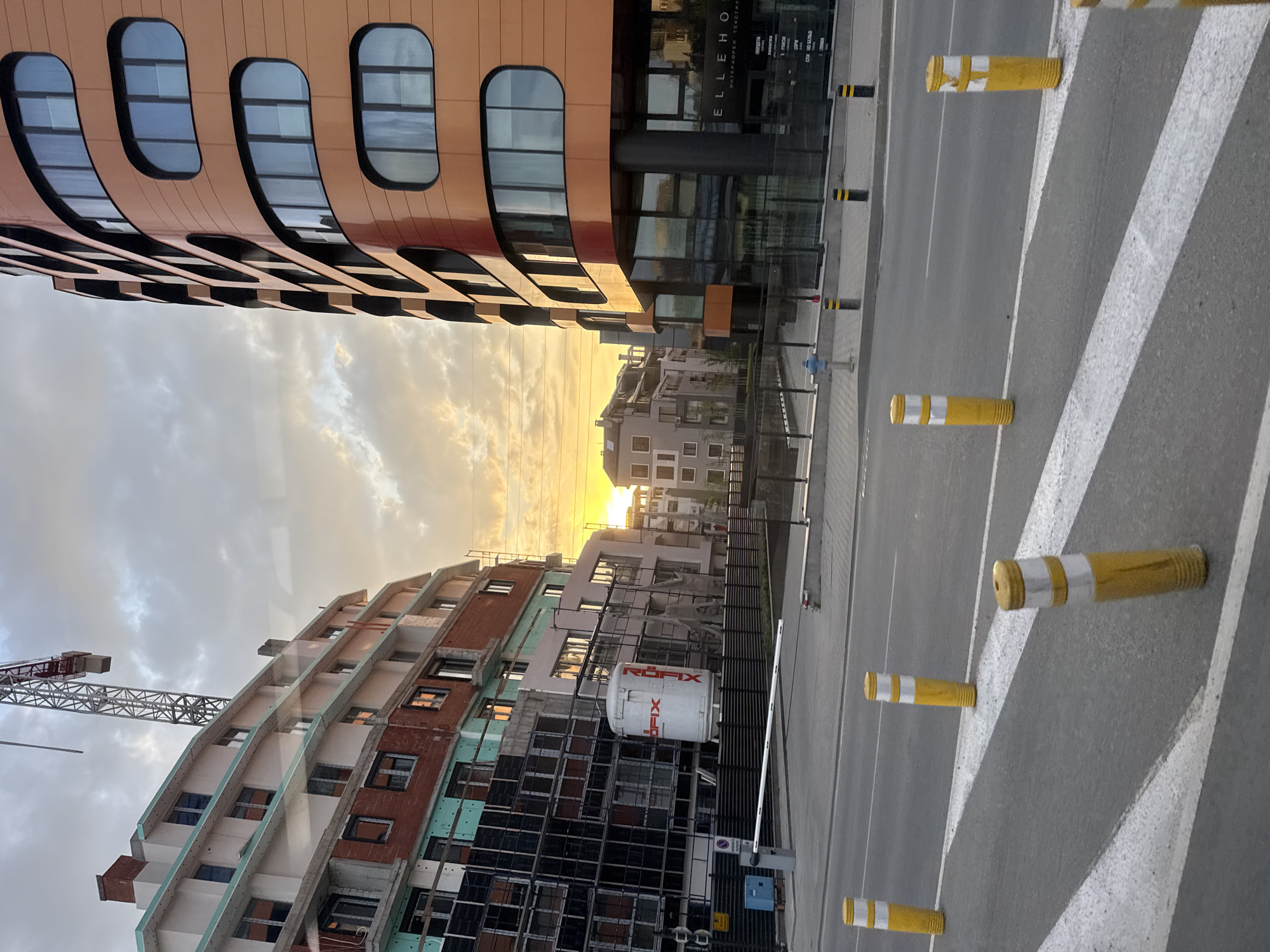 An upward-angled street-level shot shows a street in Sofia, Bulgaria, framed by two buildings against a sunset sky. On the left, a building under construction features scaffolding and a red crane. The building's facade shows exposed brick and green insulation. On the right, an orange and black building with rounded windows stands adjacent to a modern glass building. The street is marked with yellow traffic bollards and a zebra crossing. The sun sets between the buildings, casting a warm glow on the clouds.
