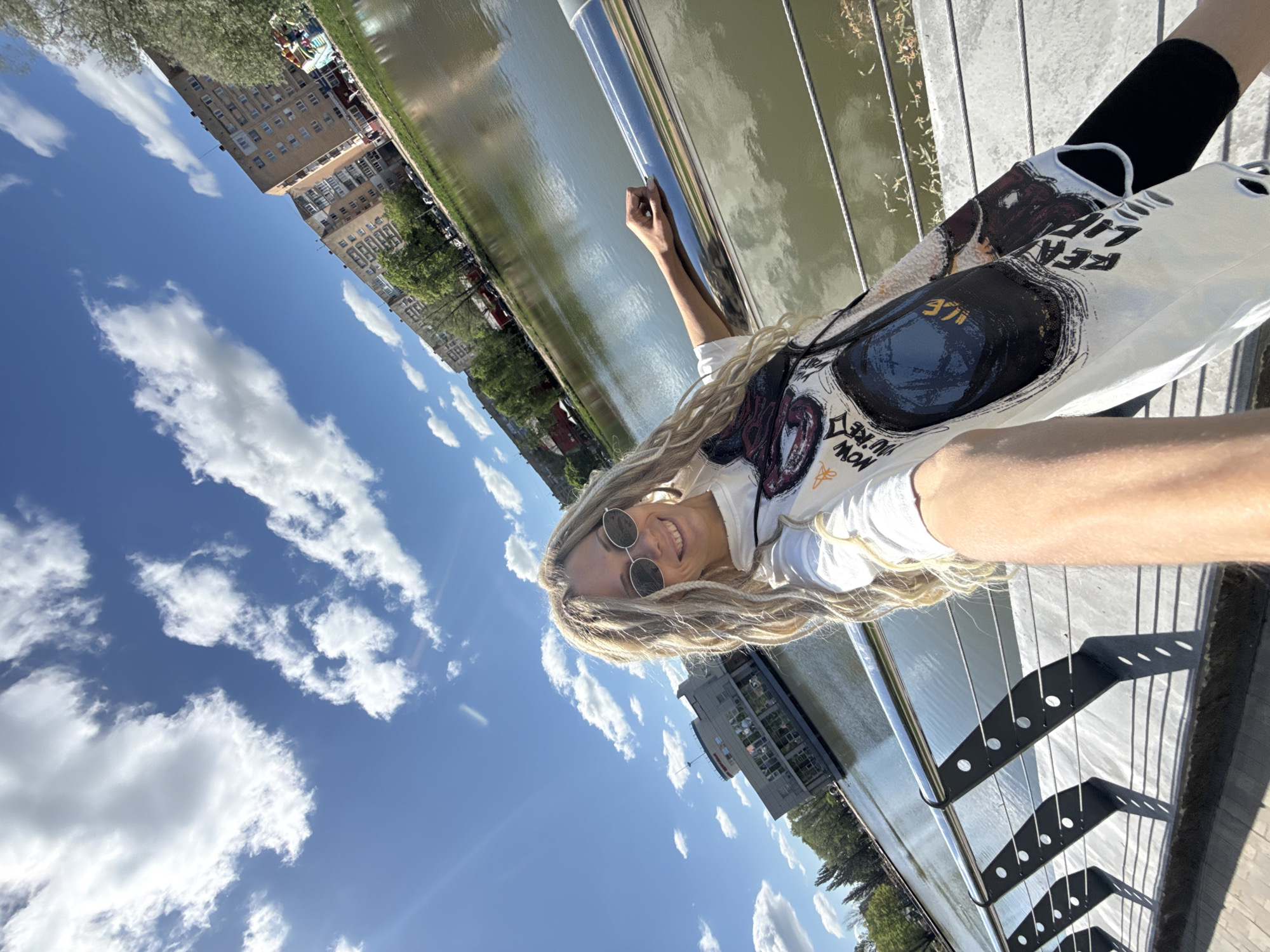 A full body selfie depicts a female wearing sunglasses, a white hoodie, and black leggings, on a walkway next to a body of water in an urban area in Unknown, Unknown. The sky is blue with puffy white clouds. In the background, buildings and trees are visible.