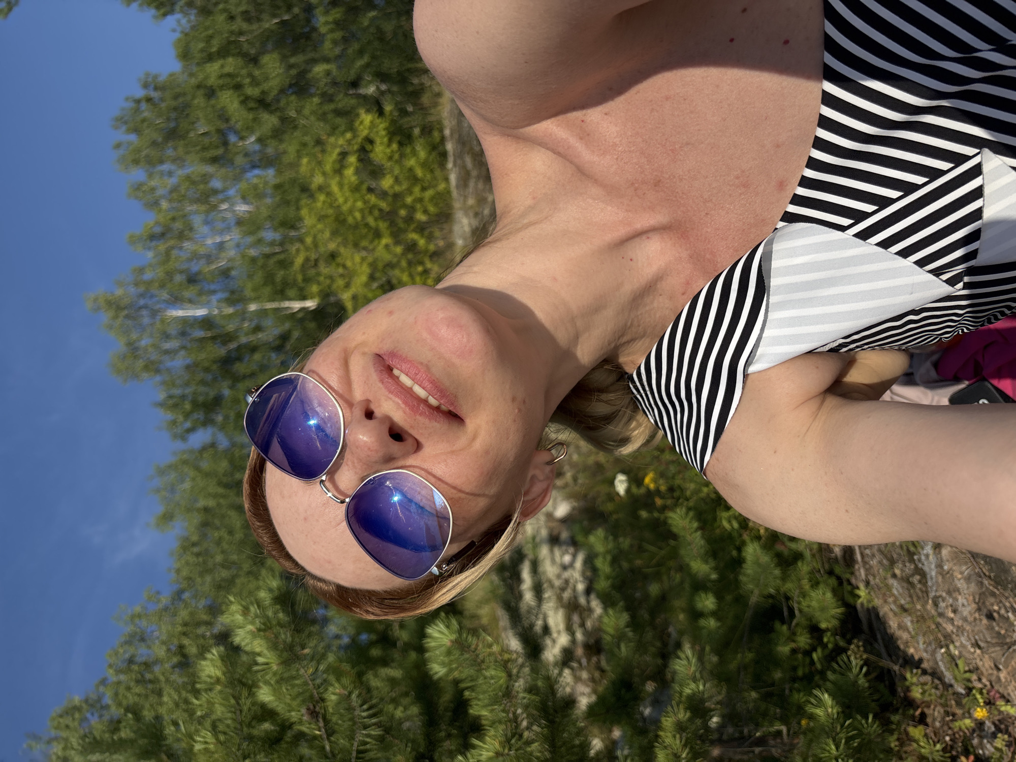 The image shows a woman wearing sunglasses and a striped swimsuit, outdoors with trees and a blue sky in the background. She appears to be taking a selfie, with her head tilted back and her mouth open in a smile. The environment suggests a natural setting, possibly a park or forest. There is no indication of the city of Shuiskaya Chupa, Russia, in the image or its context.