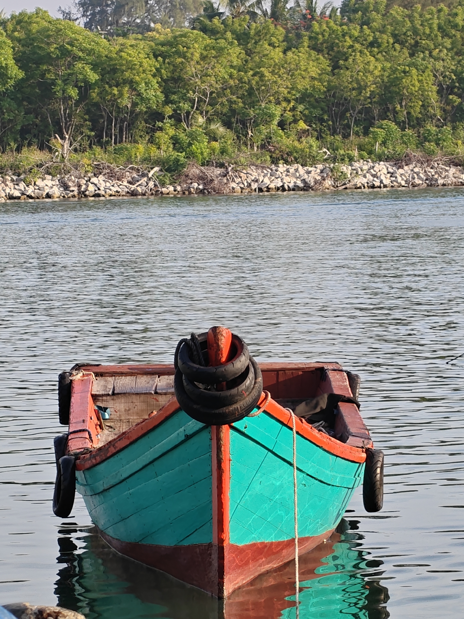 A wooden boat, painted turquoise and red, floats on calm water with a lush green forested shoreline in the background. The boat has two tires attached to its sides as fenders, and a coil of rope is visible near its bow. The shoreline consists of a rocky embankment topped with dense trees. The water reflects the sky and the boat. The scene appears to be a tranquil waterfront, possibly in the city of Lhokseumawe, Indonesia, a location known for its coastal beauty and surrounding natural landscapes.