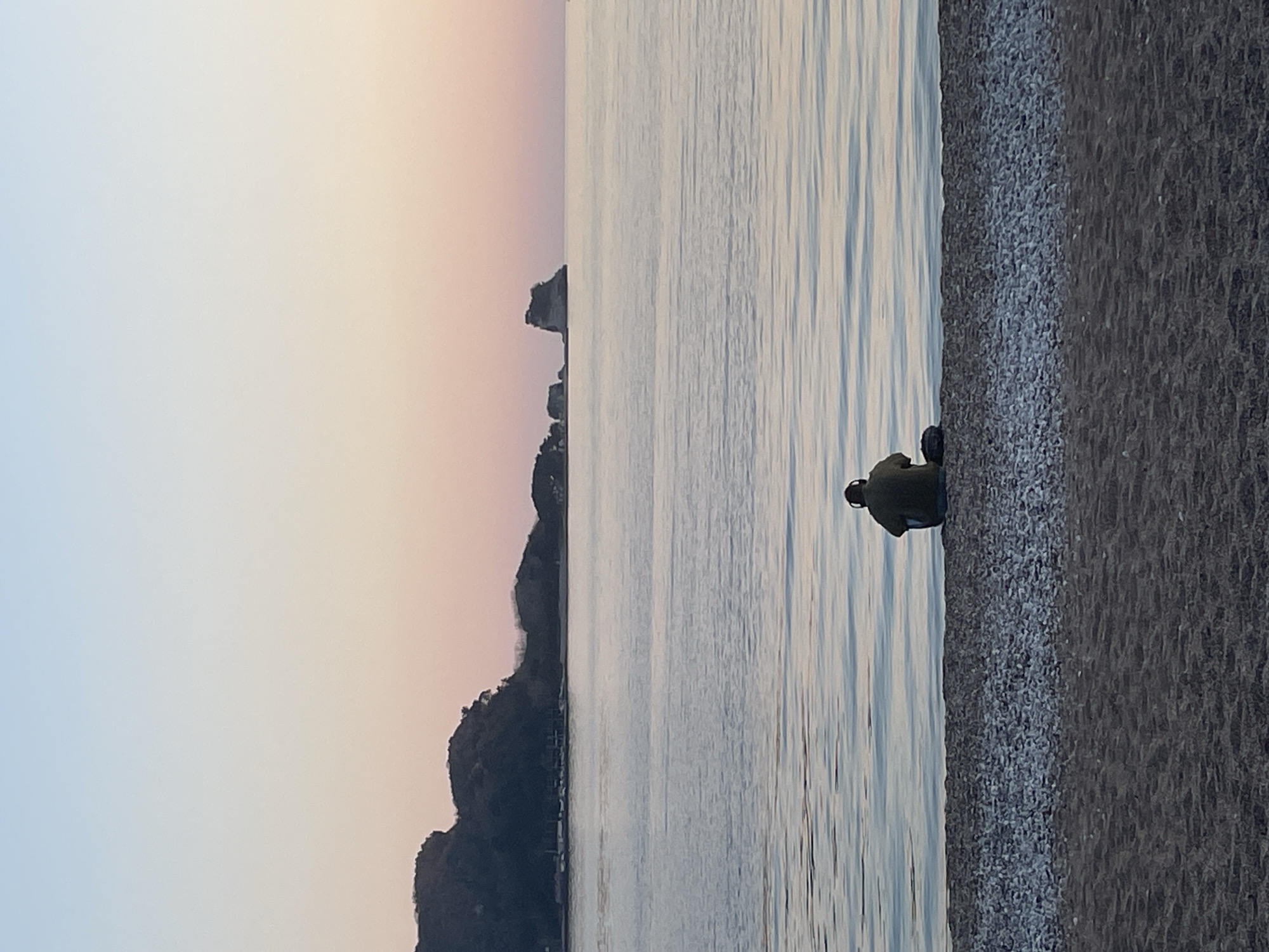 A person sits on a pebble beach at sunset with a view of the sea in Budva, Montenegro. The person is dressed in dark clothing and has their back to the viewer. The sea is calm with faint ripples. In the distance, a silhouette of rocky land protrudes from the water. The sky transitions from light blue to pink and orange as it approaches the horizon. The beach is composed of small, dark pebbles.