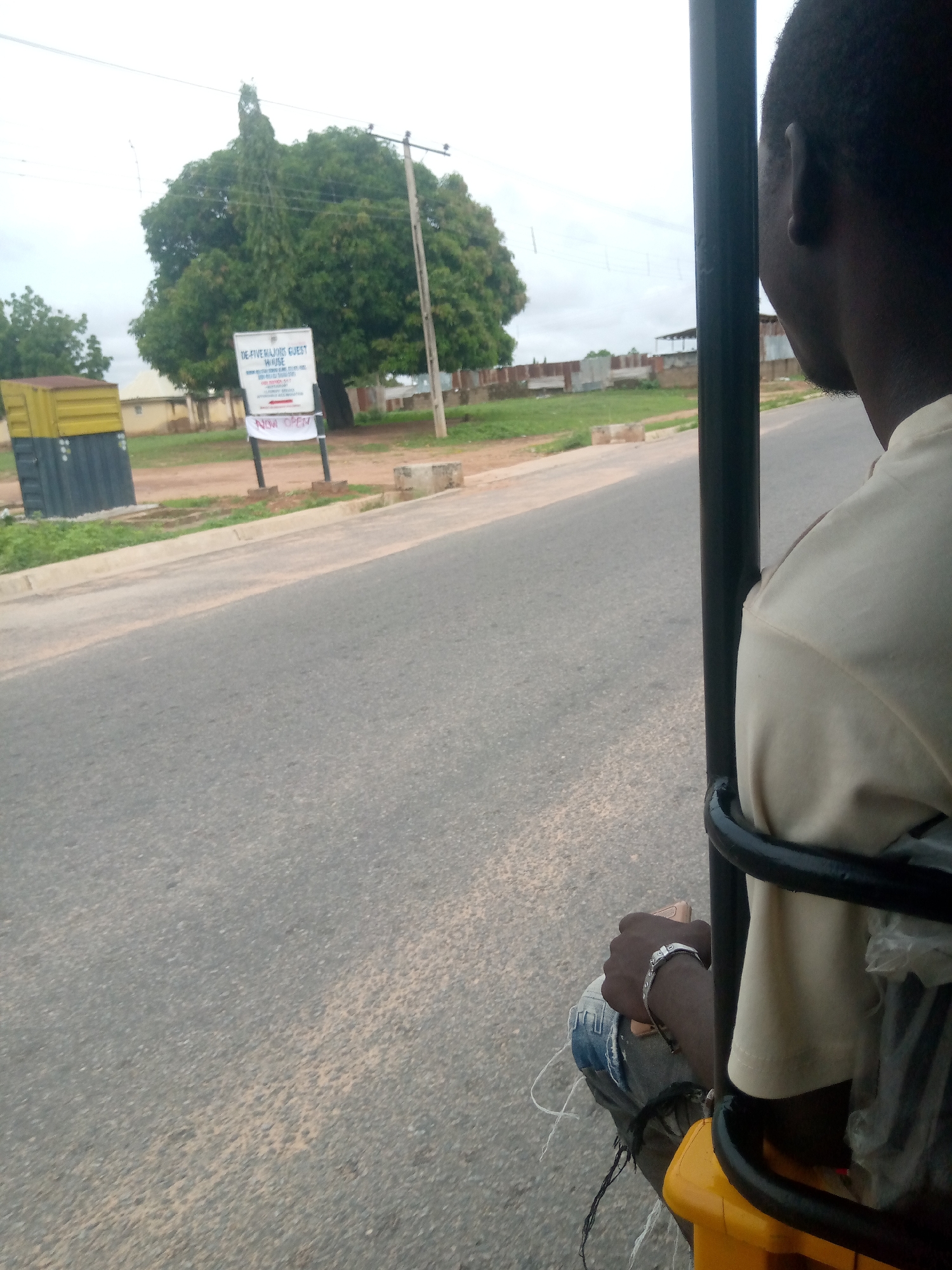 A person, viewed from behind and from the side, is seated in what appears to be the passenger area of a three-wheeled vehicle, possibly a Keke Napep. The person is wearing a light-colored shirt and blue jeans. Their left hand, adorned with a silver bracelet, rests on their lap. The vehicle is on a paved road, with the right side of the vehicle's frame visible in the foreground. To the left of the road, there is a yellow and dark gray portable structure, possibly a public toilet or a container. Further back, a large tree stands prominently, with a white signpost in front of it that reads "VE FIVE MAJORS GUEST HOUSE" and "NOW OPEN" with a red arrow pointing to the right. Beyond the tree and the sign, there are buildings and fences, suggesting an urban or semi-urban environment. The sky is overcast. The scene is consistent with a location in Jalingo, Nigeria, likely along a road with commercial establishments.