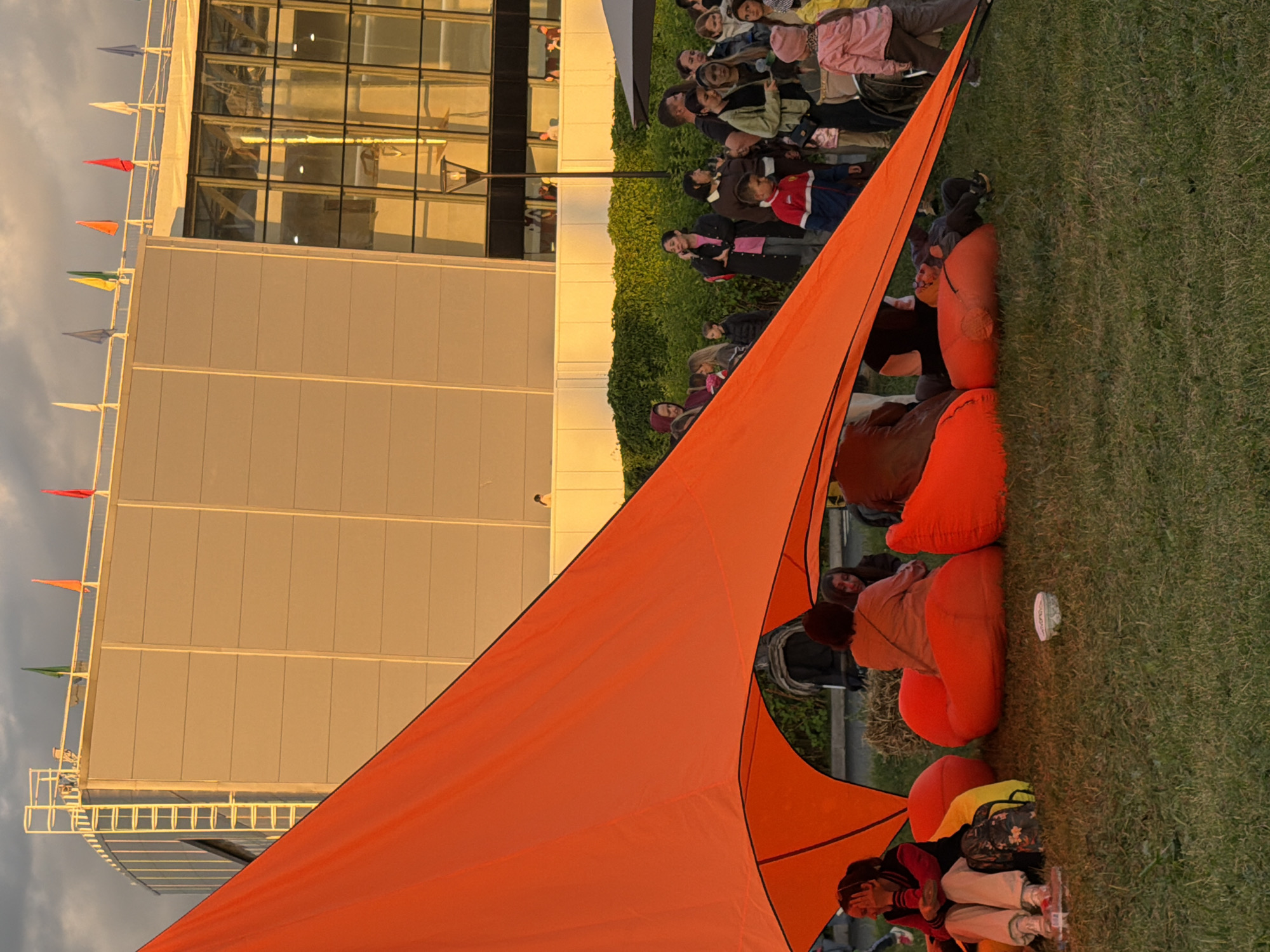 In a grassy outdoor area, a group of people are gathered under a large, bright orange canopy. Several individuals are seated on large, colorful beanbag chairs, some engaged in conversation and others looking off into the distance. The backdrop features a modern, multi-story building with large windows and a white facade. Flags in various colors are visible on a railing at the top of the building. The scene appears to be set in the late afternoon or early evening, as indicated by the warm lighting.
