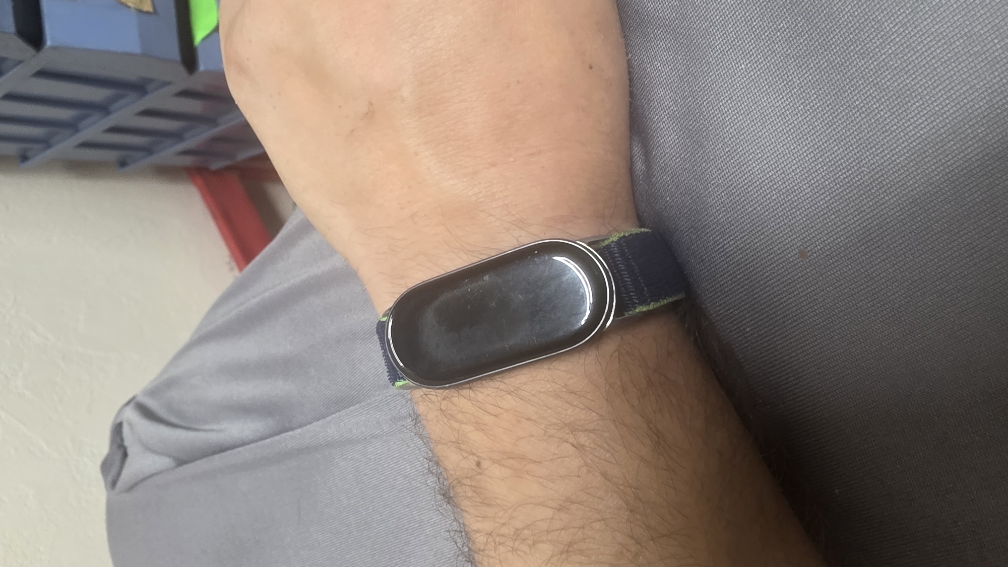 A close-up shot shows a person's left wrist, adorned with a black and green striped watchband. The watch face is a glossy black, reflecting the surroundings. The person is wearing a gray shirt, and the background features shelves filled with blue storage bins and a hint of a red shelf. The overall scene appears to be indoors.