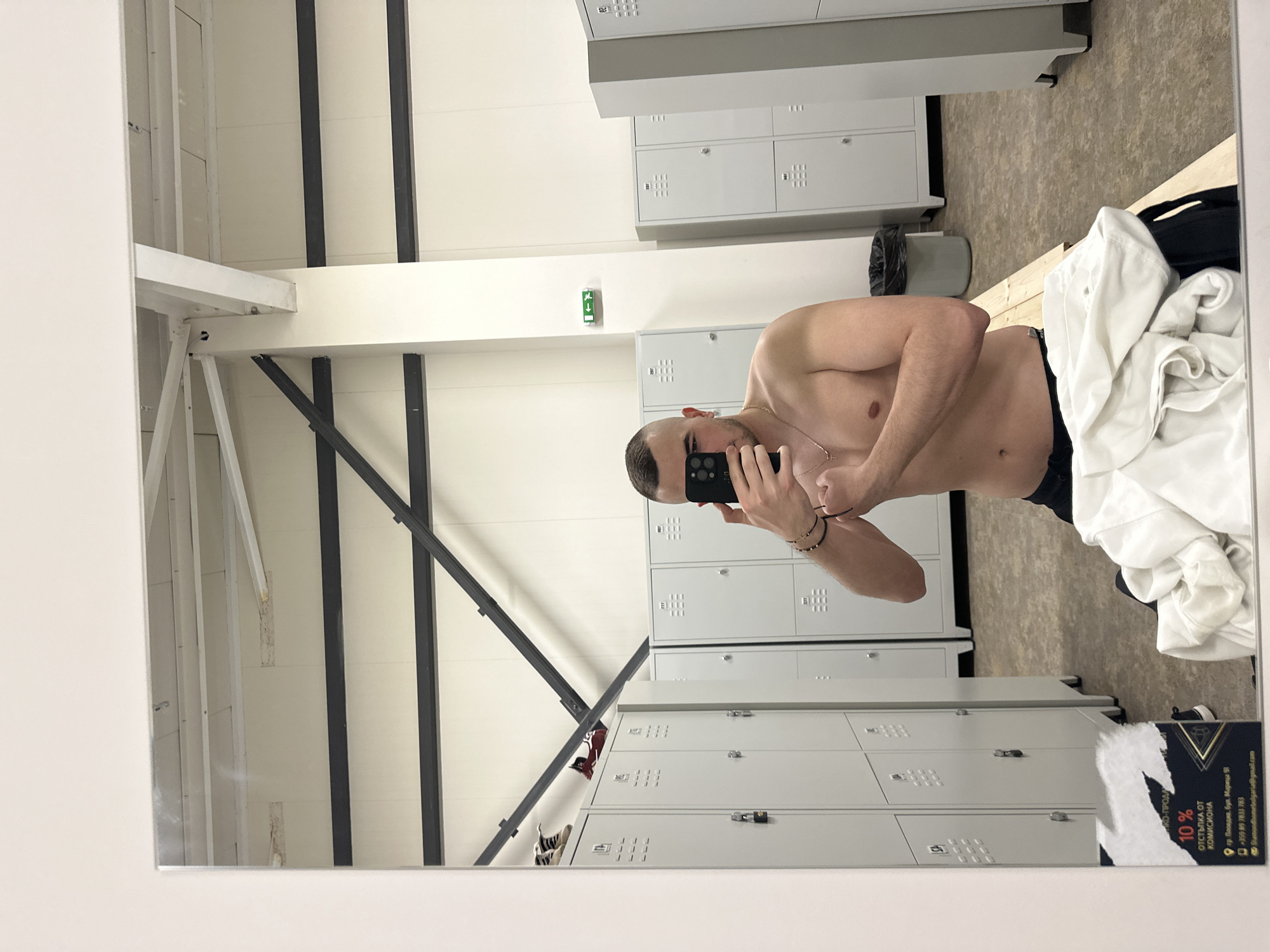 A shirtless man is taking a selfie in a locker room. He is holding a phone in front of his face, obscuring most of his features except for his forehead and short hair. He wears a gold chain around his neck and bracelets on his wrist. The locker room has rows of gray lockers and a concrete floor. There is a pile of white clothing on a bench next to the lockers. The reflection in the mirror also reveals shoes and the structural supports of the building.
In the lower right corner of the image, a business card is visible, advertising a 10% discount from a commission at "ПКО-ПРОДА" located at 91 Maritsa Blvd, Plovdiv, Bulgaria.