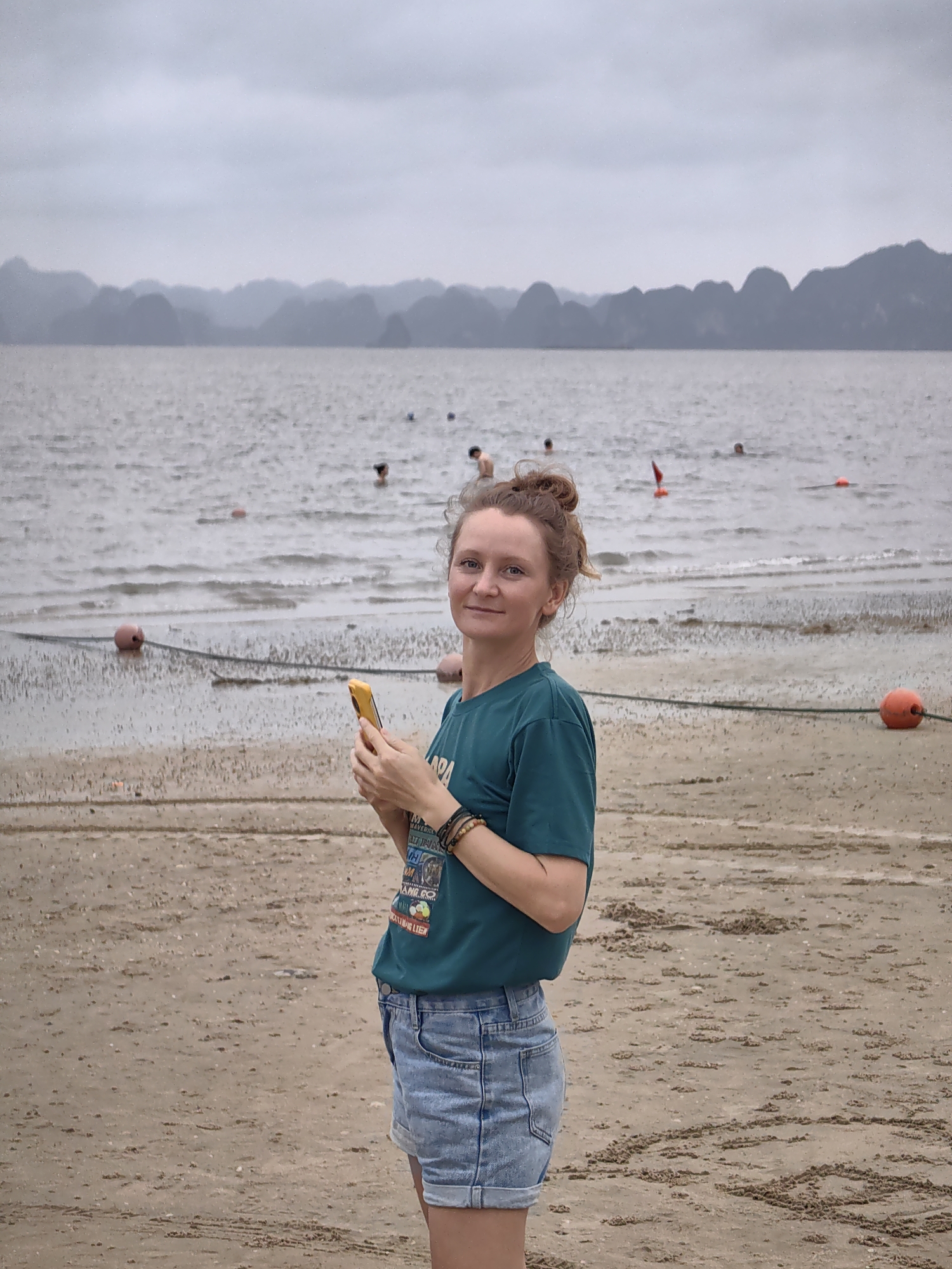 The image features a woman standing on a sandy beach, looking directly at the camera with a soft smile. She has light skin and reddish-brown hair tied in a bun. She is wearing a dark teal t-shirt, which has partially visible text including "MAVERICKS" and other graphics, and light blue denim shorts. Multiple beaded and thread bracelets adorn her right wrist, and she holds a bright yellow smartphone in her hands.
Behind her, the calm, greyish sea extends towards a distant horizon. Several people are visible swimming or wading in the water, appearing as heads and shoulders above the surface. A dark rope with orange buoys stretches across the water closer to the shore, likely marking a swimming area. The sandy beach in the foreground displays patterns and small mounds, suggesting tidal action.
In the far background, a distinctive chain of dark, jagged karst limestone mountains rises from the water, silhouetted against a uniformly overcast, grey sky. The scene is set on an apparently cloudy day. The presence of the karst mountains, combined with the provided context, indicates the location is Bai Chay Ward, Vietnam. The overall atmosphere is tranquil, with people enjoying the water despite the overcast weather.