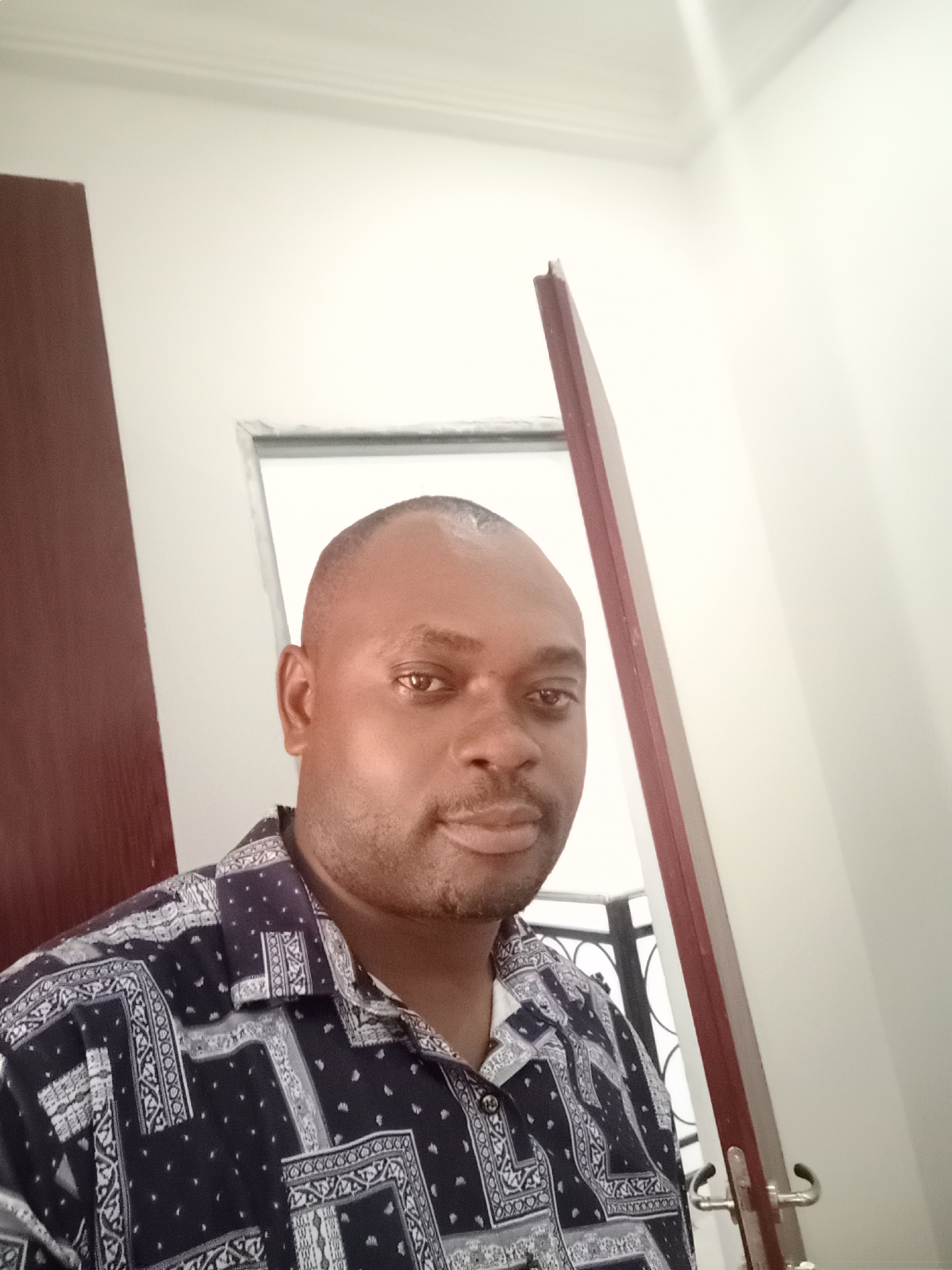The image is a selfie taken indoors, likely in a residential area of Gwarinpa, Nigeria. The person in the image is a middle-aged Black man with a close-cropped haircut, wearing a patterned, dark blue and white collared shirt. He has a mustache and a slight beard. The background shows a partially open doorway. The door frame is dark wood. Behind the partially open door, a window with dark bars is visible. The walls are painted a light color, likely white or off-white. A door handle is visible on the right side of the frame.