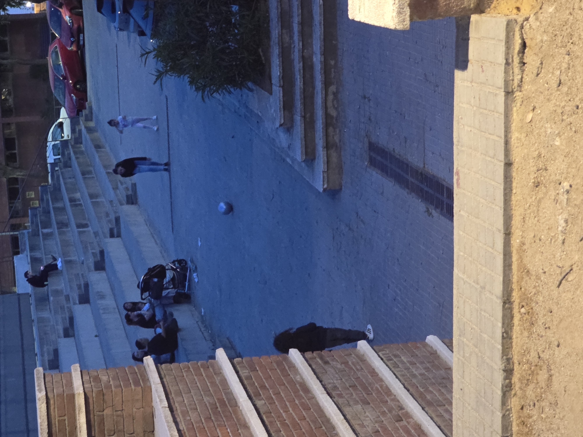 The image shows an outdoor scene at dusk in Cornellà de Llobregat, Spain, with a view from an elevated position overlooking a tiered, open-air seating area or amphitheater. Several individuals are scattered throughout the seating area, some sitting, others lying down, and a few walking on the path above. Objects such as bags and a stroller are visible among the people. Above the seating area are parked cars and some vegetation. The foreground includes the edge of a brick structure and a concrete pillar. The overall scene is bathed in a dim, blueish light.