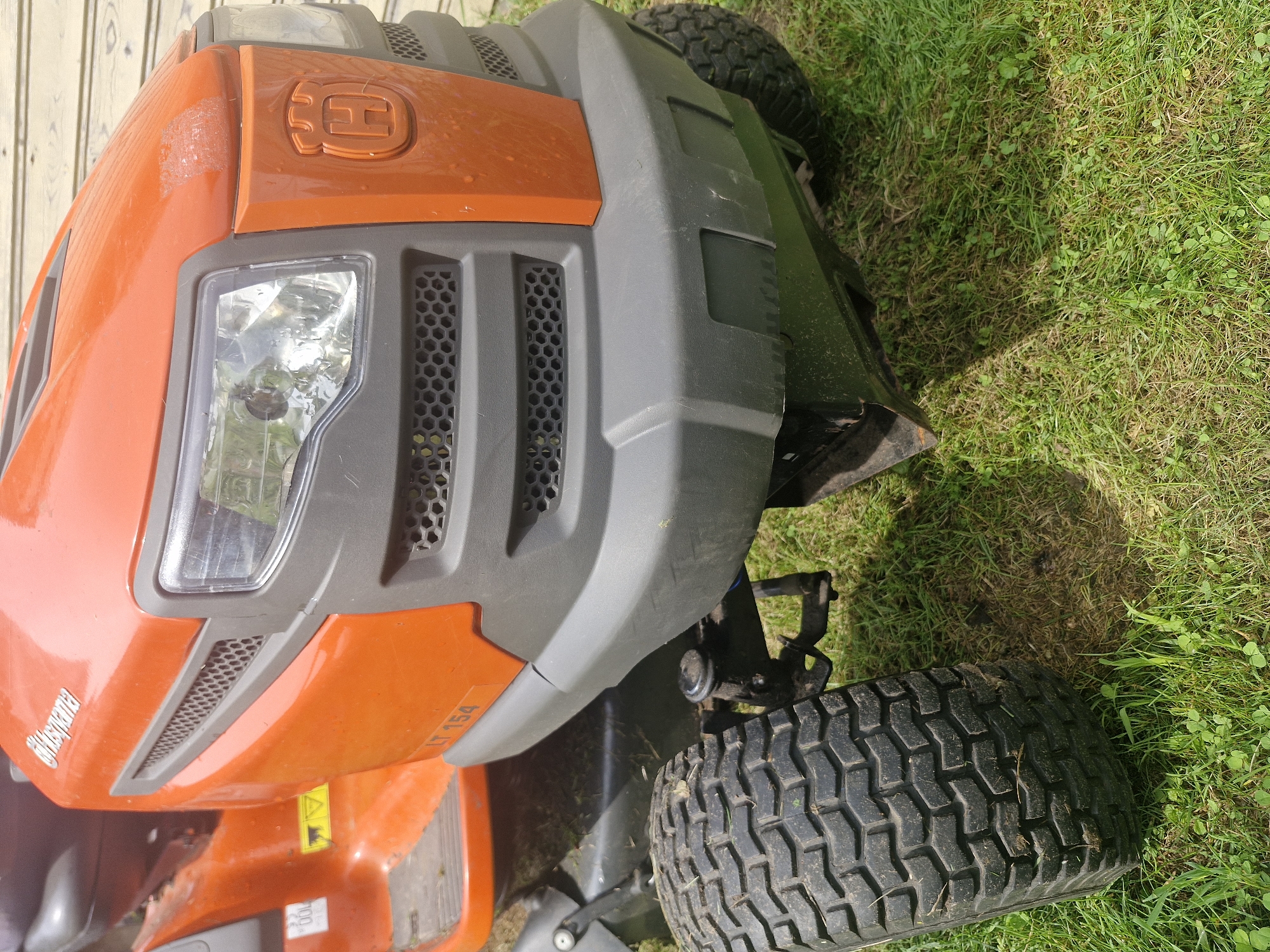 A close-up, eye-level, full shot captures the front of an orange and gray Husqvarna LT 154 riding lawn mower parked on a patch of green grass and dirt, likely in a residential yard in Ķekava, Latvia. The mower's prominent orange hood features a silver Husqvarna logo on the upper left and a recessed gray grille with a hexagonal pattern. The headlights are large and clear, and the gray bumper below has a textured, hexagonal pattern as well. The mower's front wheel, with a rugged tread, is visible on the left side of the frame. The grass is a mix of green blades and some dry, brown patches. The background shows a wooden wall, suggesting the mower is near a building.