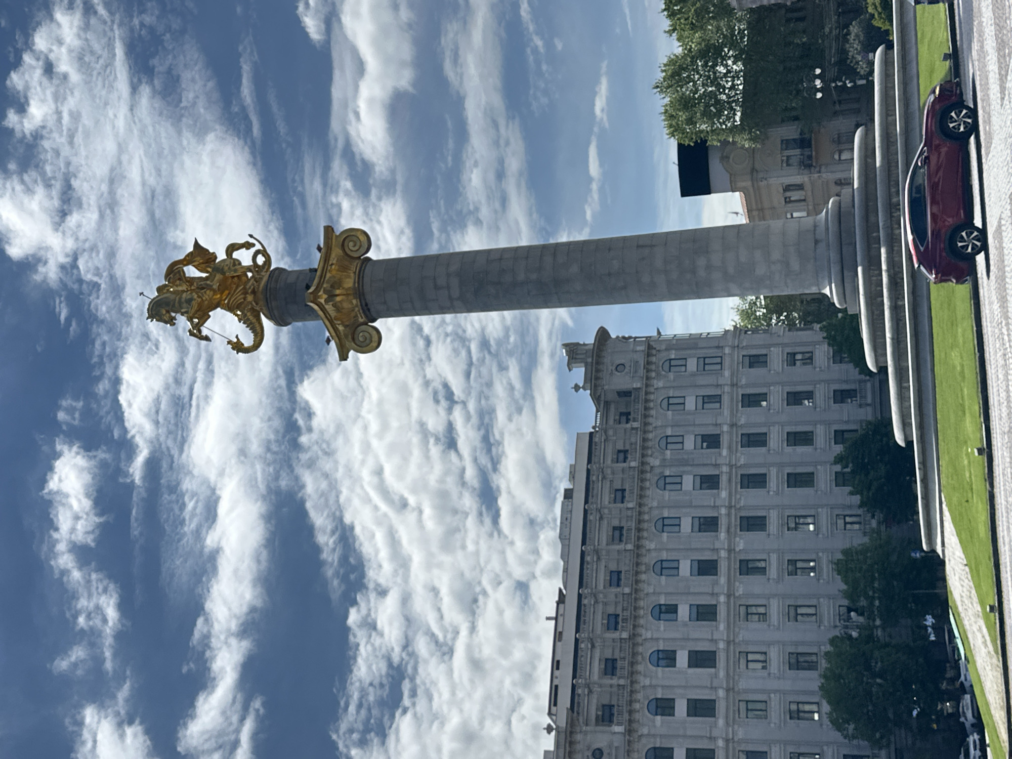 The image shows the St. George's Monument, located in Tbilisi, Georgia. The monument features a tall column topped with a golden statue of St. George slaying a dragon. The sky is blue with scattered white clouds. A large, grey building with many windows is visible in the background, along with trees and a portion of a red car. The base of the monument consists of multiple layered rings.