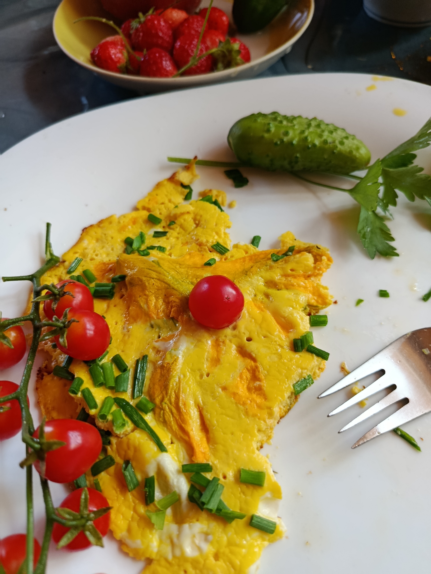 A close-up, overhead shot shows a partially eaten omelet on a white plate. The omelet is a bright yellow, sprinkled with chopped green chives. A cluster of cherry tomatoes on a vine is positioned to the left of the omelet, with one tomato resting on top of it. To the right of the omelet, a silver fork rests on the plate, and a small, bumpy cucumber and a sprig of parsley are visible in the background, suggesting a meal in a home setting. In the upper portion of the image, a bowl of strawberries can be seen. The lighting is bright, creating a well-lit scene.