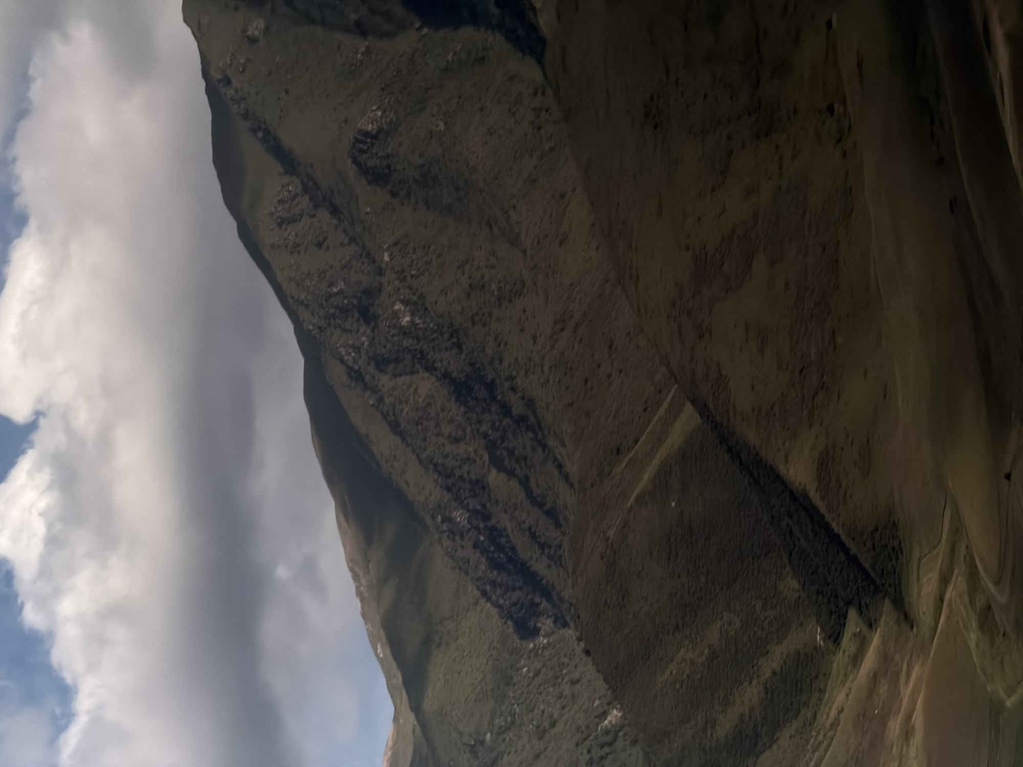 The image depicts a dramatic, tilted view of a mountainous landscape, likely Karagai Bulak, Kyrgyzstan. The dominant subject is the rugged terrain, characterized by steep, dark slopes covered in vegetation. These slopes are crisscrossed by deep shadows, suggesting a time of day when the sun is casting long shadows, perhaps late afternoon or early morning. The texture of the ground appears rough and uneven, with scattered rocks and patches of earth.
A striking feature of the scene is the contrast between the dark mountainside and the bright, cloudy sky. Large, white clouds dominate a significant portion of the frame, with glimpses of blue sky peeking through. This suggests dynamic weather conditions, possibly a partly cloudy day. The clouds are illuminated from above, further emphasizing the time of day and the presence of sunlight.
There are no discernible people or man-made objects in the frame. The focus is entirely on the natural, imposing grandeur of the mountains. The overall mood conveyed is one of wild, untamed nature. The extreme tilt of the camera adds a sense of disorientation or a unique perspective, as if the viewer is looking up at the mountain from a precarious position or experiencing a moment of dramatic ascent or descent. No visible text is present in the image.