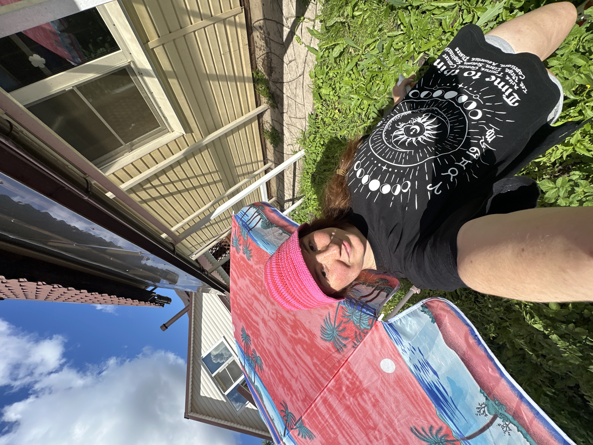 A young woman with freckles and a pink knitted hat is lying in the grass, taking a selfie under a red and blue beach umbrella. She is wearing a black t-shirt with a celestial design and the words "Time to Shine" printed on it. The umbrella has a tropical beach scene depicted on it. In the background, there is a light-colored house with a brown roof and a partially visible window with a reflection of the umbrella. The house appears to be made of siding. The scene is outdoors, with green grass and plants surrounding the woman. The sky is blue with white clouds. The location is the city of Nizhny Novgorod, Russia, likely a backyard or garden area.