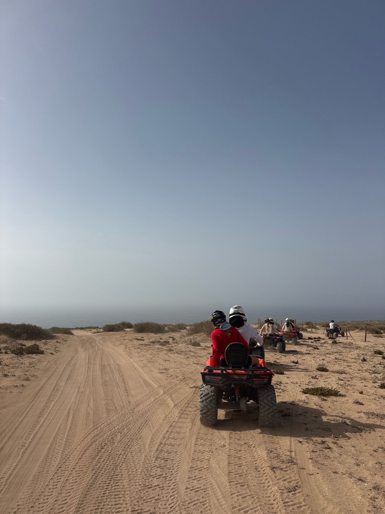In Tamri, Morocco, a group of individuals is seen riding quad bikes along a sandy track that leads towards the ocean. The sky is a clear, light blue, with a faint haze obscuring the horizon where the sea meets the sky. The terrain is predominantly sandy with sparse, dry vegetation dotting the landscape. The quad bikes are red, and the riders are wearing helmets and protective gear. The scene captures a moment of outdoor recreation in a coastal desert environment.