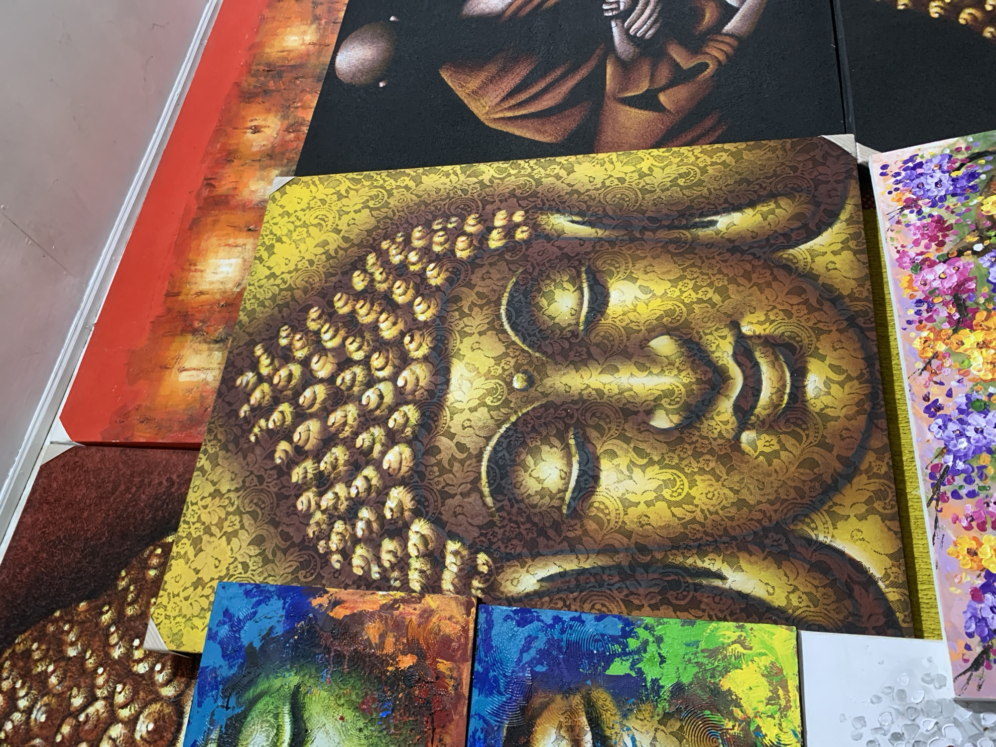 A close-up, high-angle shot depicts a collection of paintings, possibly displayed for sale in Ubud District, Indonesia. The primary subject is a rectangular painting of a golden Buddha head with intricate details, overlaid with a lace-like pattern. The head is oriented sideways, with the top of the head towards the left. Surrounding this central painting are sections of other artworks, including a painting with red and orange tones, a dark painting of a figure, and one with vibrant purple and pink floral patterns. A few additional abstract paintings with green and blue tones are visible at the bottom of the frame. The composition suggests an indoor setting, possibly a shop or gallery. The environment is somewhat cluttered with overlapping paintings, indicative of a commercial art display.