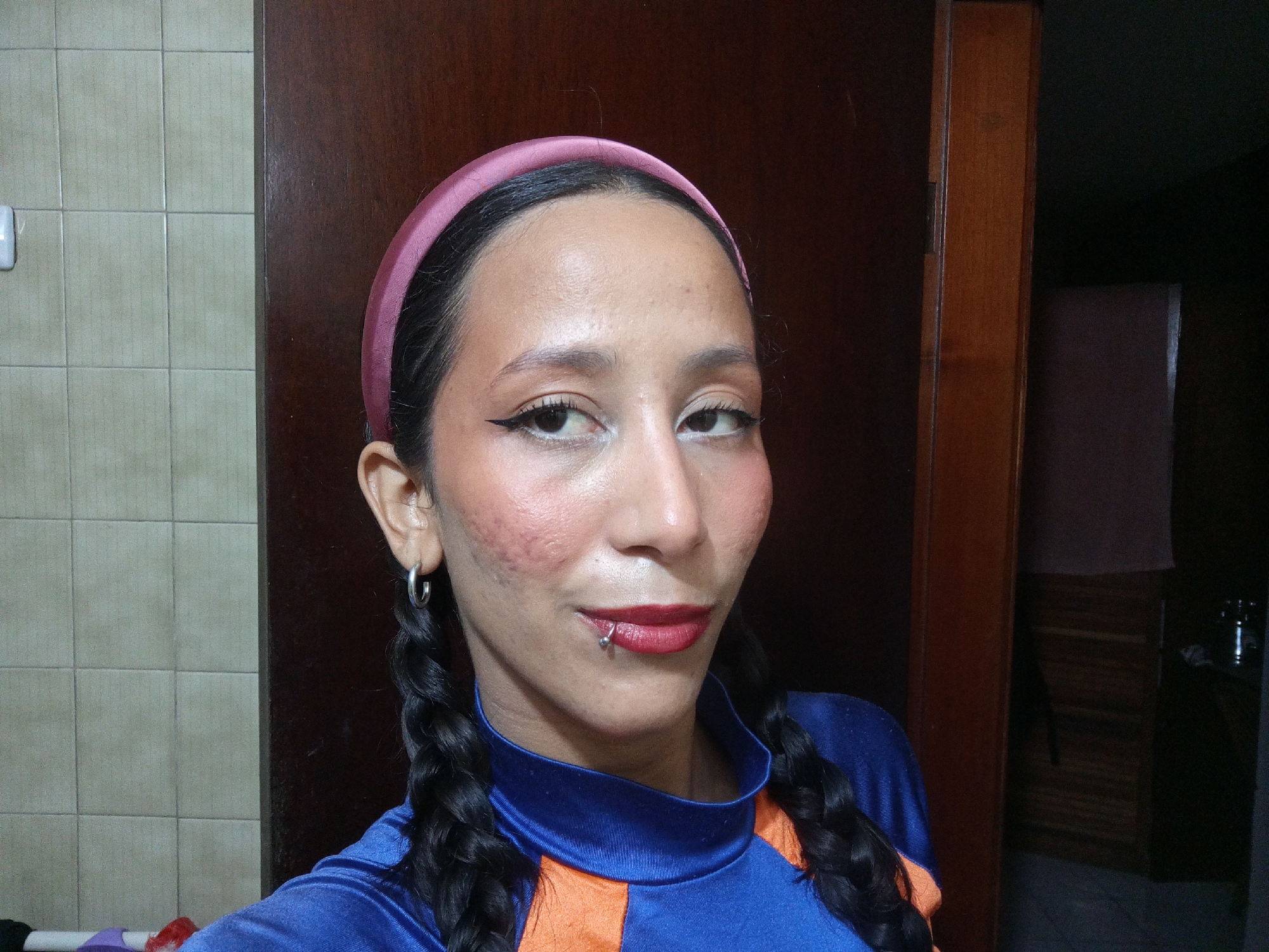 A person is centered in the frame, facing the camera, wearing a pink headband, with black braided pigtails. They have a lip piercing, dark eyeliner, and red lipstick. The person is wearing a blue and orange top. Behind the person, to the left, is a wall with beige square tiles. To the right is a dark brown wooden door. The location is not discernible from the provided image.