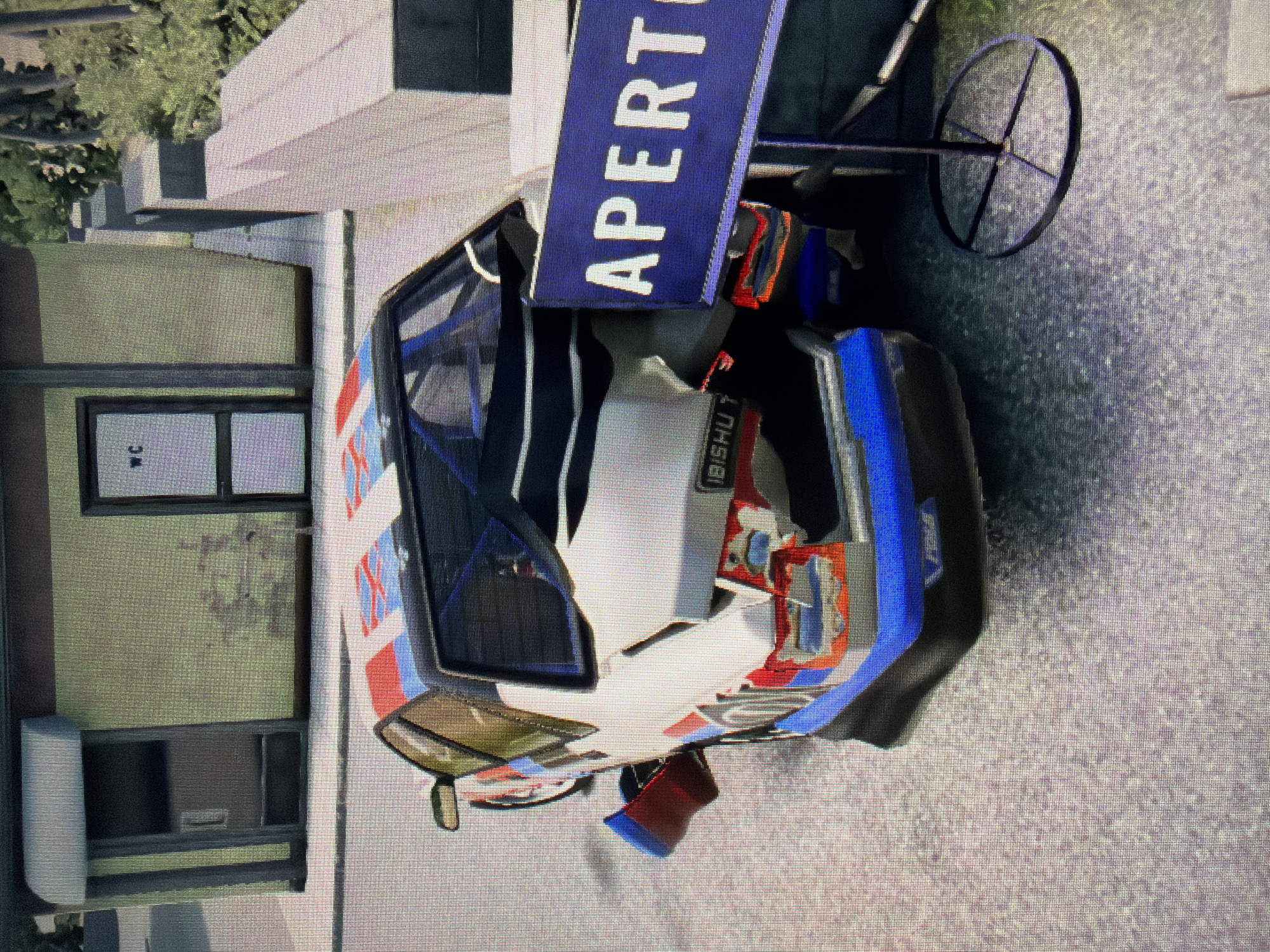 The image displays a white, blue, and red customized car, likely a rally or race car, stationary next to a building on a paved surface. The car shows visible damage, particularly to its front bumper and rear, with some internal components like a roll cage visible. Partial text "BISHU T" can be seen on the car's side.
The building, situated to the left of the car, features light-colored, textured walls, dark window frames, and a door with a clear "WC" sign, indicating a public restroom facility. Green foliage is visible in the background beyond the building.
Behind the car, a prominent blue sign with the white capitalized text "APERTO" (Italian for "Open") is mounted on a stand that includes a spoked wheel. The ground beneath the car and building is a light gray, possibly concrete or asphalt. The scene is outdoors under bright daylight. The pixelated quality of the image suggests it may be a screenshot from a video game.