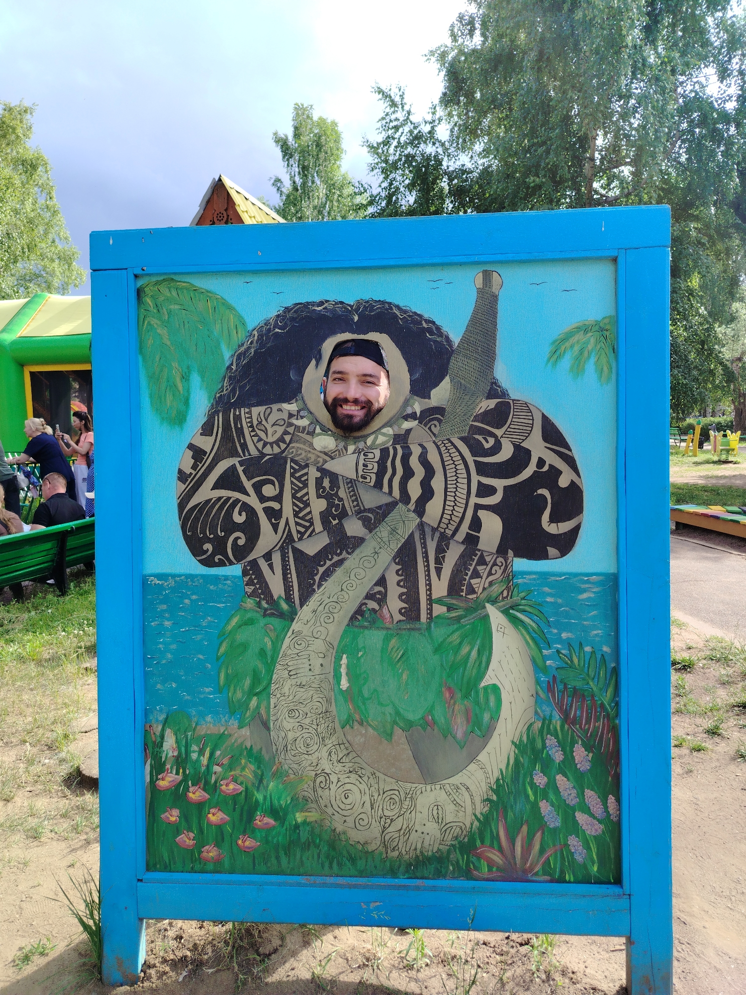 A man is posing for a photograph in a cut-out board featuring a painted character with Polynesian-style tattoos, positioned in front of a body of water. He is wearing a black beanie and a black t-shirt. The board is blue and is located outdoors, with trees and a playground visible in the background. It appears to be a sunny day. The scene is in the city of Chaikovsky, Russia.