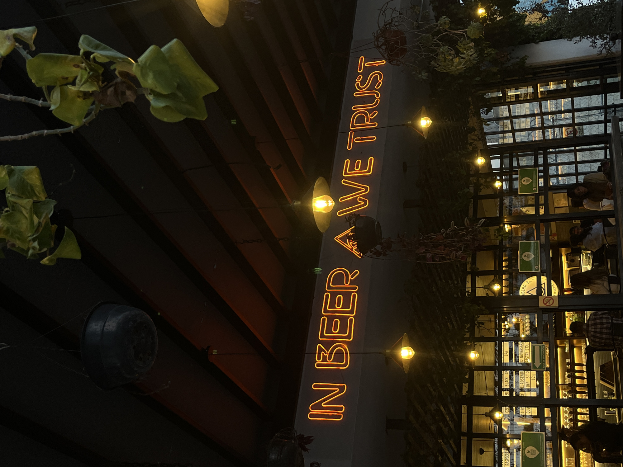 The image depicts an outdoor area in San Andrés Cholula, Mexico, likely a bar or restaurant, at night. A neon sign that reads "IN BEER WE TRUST" is prominently displayed. Several hanging lights are visible, providing illumination. In the background, there are visible people and what appears to be a well-stocked bar through a window or open doorway. Plants, including hanging foliage and potted plants, are incorporated into the decor. The overall setting has a relaxed, inviting atmosphere. A decorative flowerpot hangs upside down at the left of the image.