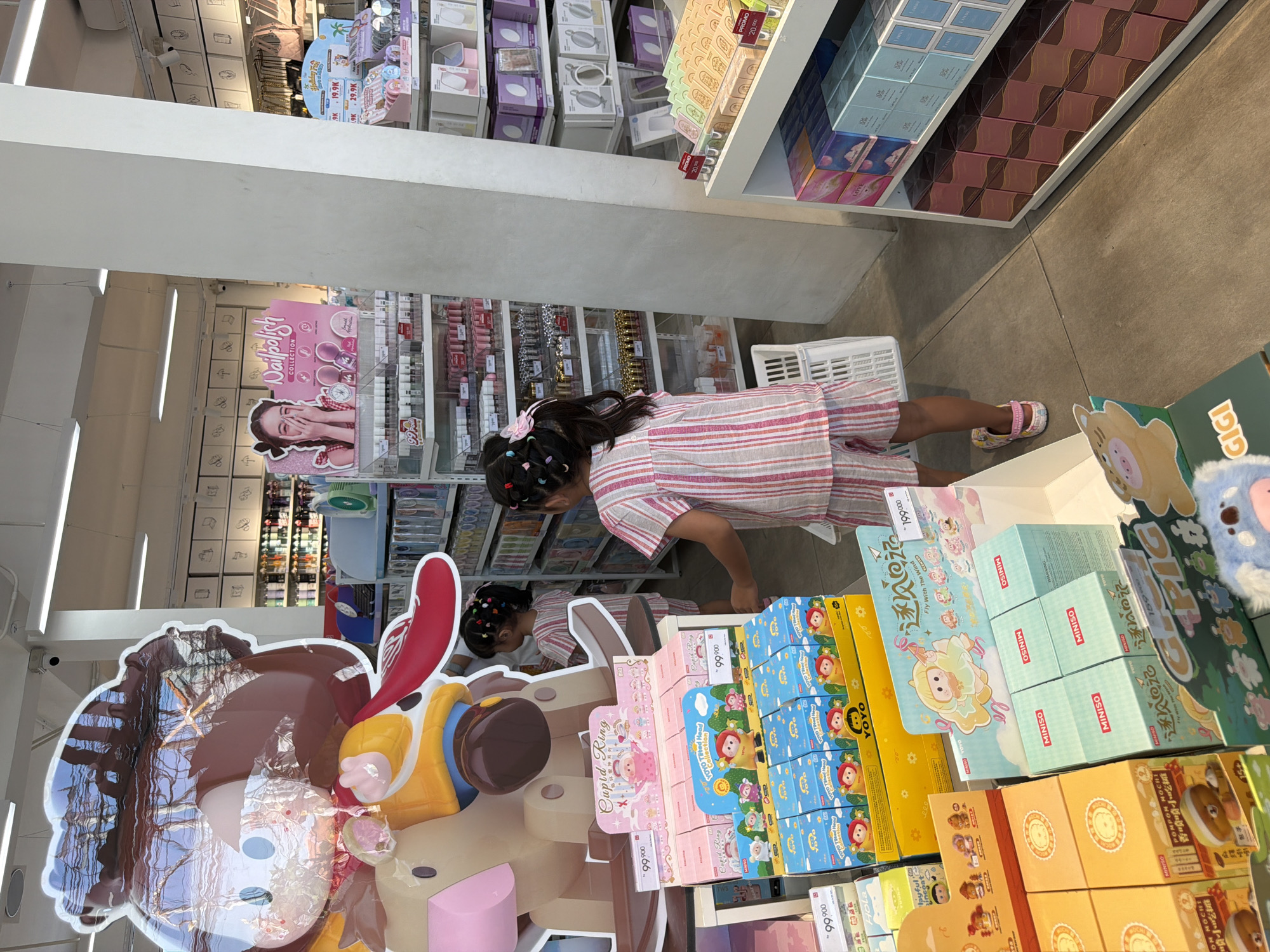 In a brightly lit retail store, a young girl with dark hair tied with a pink bow is looking at shelves filled with various products. She is wearing a pink and white striped dress and sandals. She is bending over, appearing focused on the items on display. Behind her, more shelves are visible, stocked with boxes of cosmetics and other merchandise. A large, colorful cutout of a character, possibly a toy or mascot, stands to the left of the frame. The store appears to be a boutique or a section within a larger store, possibly selling beauty products or gifts. The overall atmosphere is clean and organized, with a focus on colorful packaging. Visible text on the products includes "MINISO," "YOYO," "Cupid Ring," "Nailpolish COLLECTION," and prices like "99.900." The setting suggests it's daytime, and the environment is indoors.