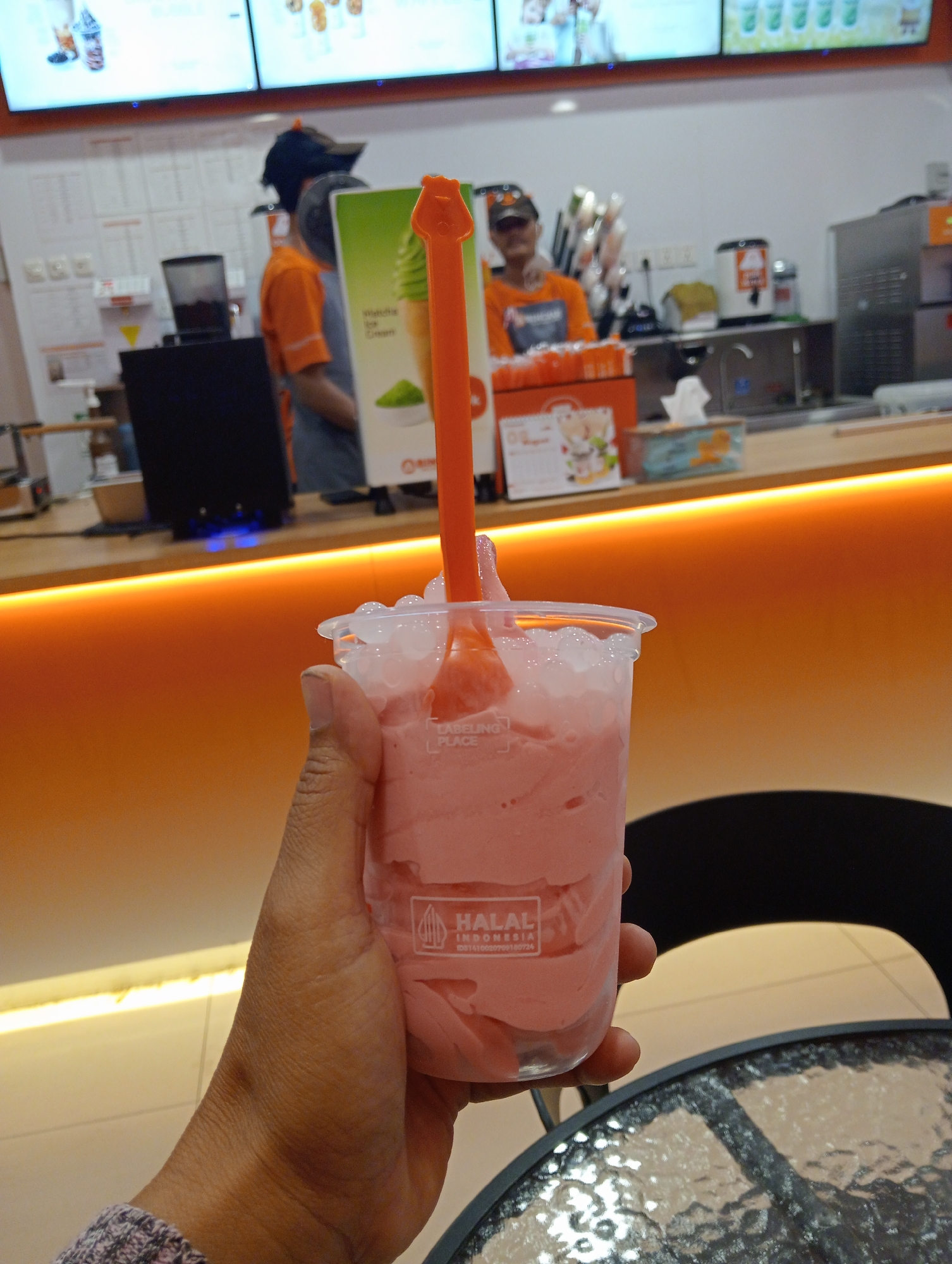 The image shows a person holding a pink smoothie in a clear plastic cup with a plastic spoon. The smoothie is topped with crushed ice and some small white spheres. The cup has a label that reads "HALAL INDONESIA" with some text below it. In the background, there is a counter with staff members, display screens showing menus, and a drink-making station. The setting appears to be a cafe or a similar establishment. The orange lighting in the foreground suggests this is an indoor commercial space. The overall scene is a close-up of a person enjoying a drink.