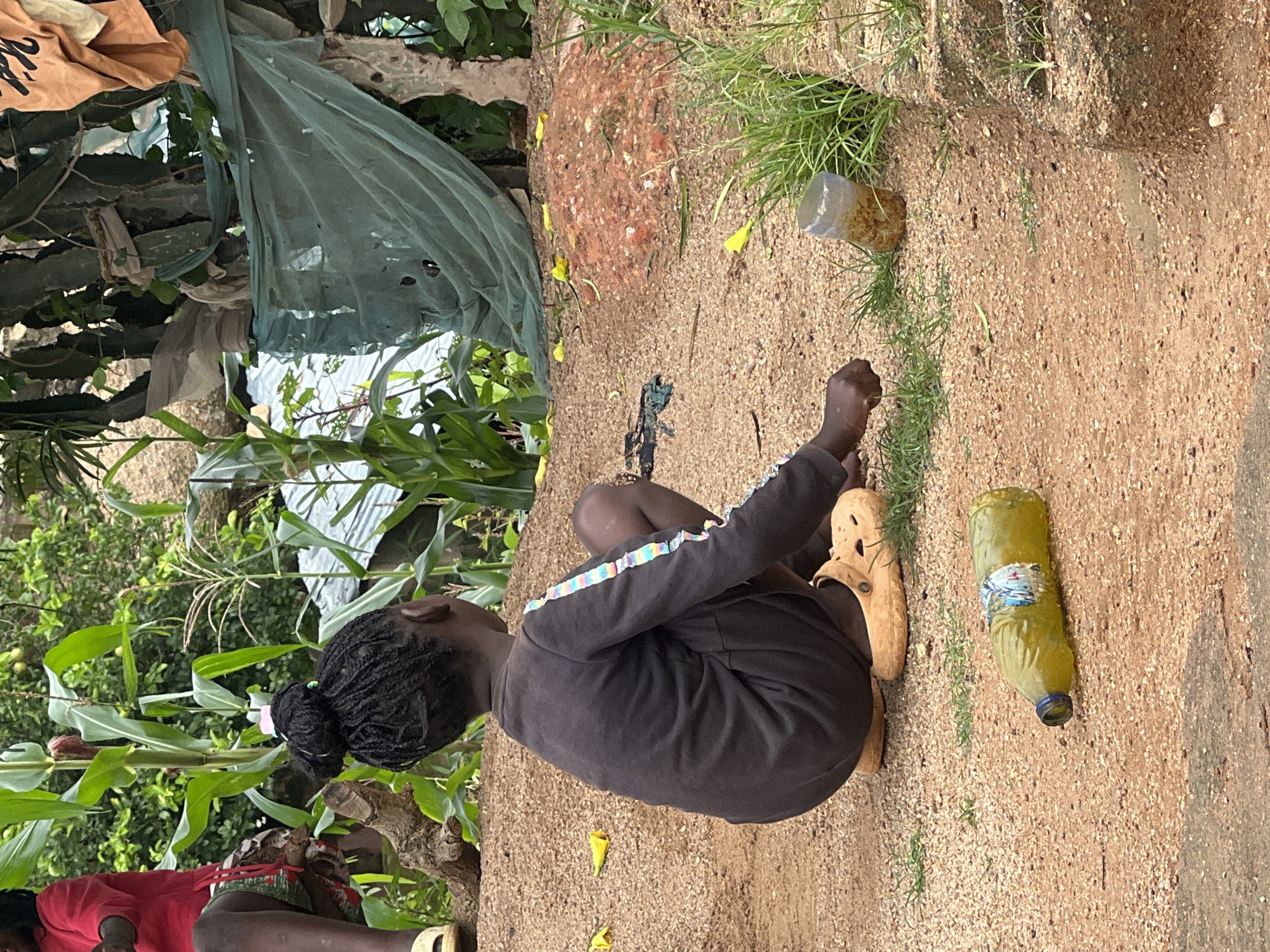 In Jos, Nigeria, a young Black girl with braided hair in a bun is seen from behind, squatting on a sandy dirt path. She is wearing a dark long-sleeved shirt with colorful stripes on the sleeves and tan slip-on shoes. In front of her, to her left, is a partially filled green bottle lying on its side. To her right, further away, is a clear plastic cup containing a brown substance and some green grass growing around it. The environment appears to be an outdoor setting with lush green corn stalks and trees in the background, some of which are covered with a green mesh cloth. Another child, wearing a red shirt, is visible in the upper left corner of the frame. The ground is a mixture of sand and small pebbles, with scattered yellow flower petals.