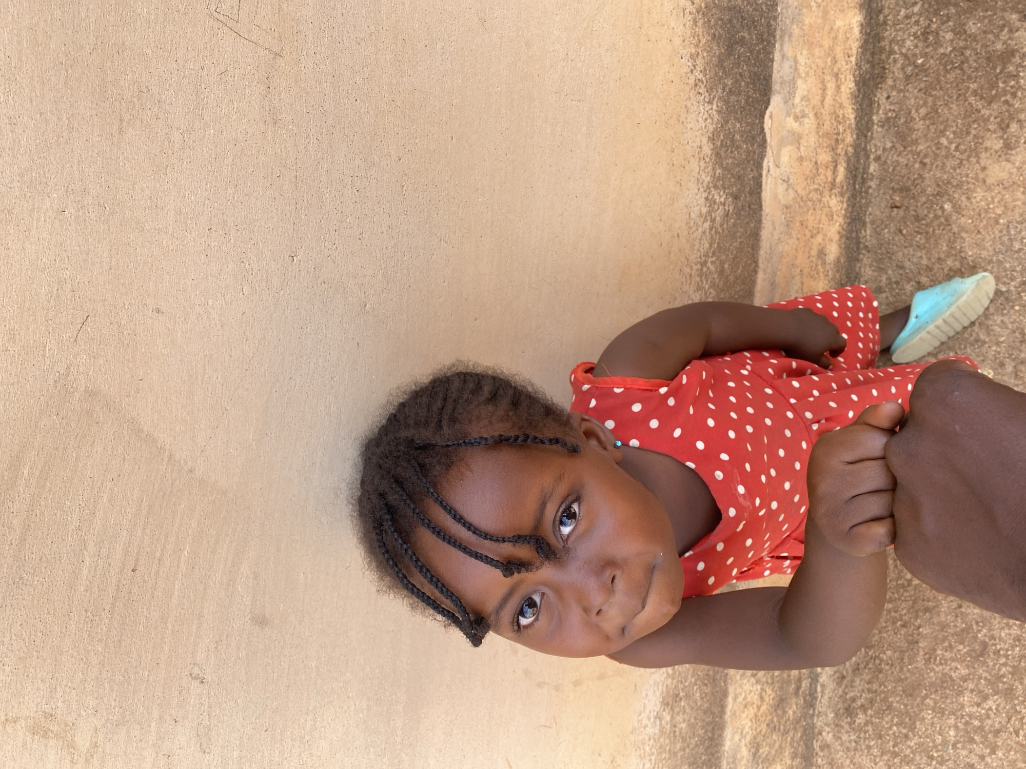 The image captures a close-up, angled view of a young child, likely a girl, in what appears to be an outdoor setting in Kuru Dungus, Nigeria. The child has dark skin and short, braided hair. She is dressed in a red sleeveless dress with white polka dots. Her eyes, dark and observant, are directed towards the viewer, conveying a sense of innocent curiosity.
Her right hand is clasped around a larger, dark-skinned adult hand or fist, suggesting a secure or comforting grip. A light blue shoe or sandal is visible on her left foot, resting on the ground.
The setting consists of a light brown, textured wall on the left side of the frame and a rough, darker brown ground surface on the right. The wall shows some minor imperfections and discolouration, while the ground surface could be packed earth or worn concrete. The bright, natural light suggests it is daytime. There is no visible text in the image.