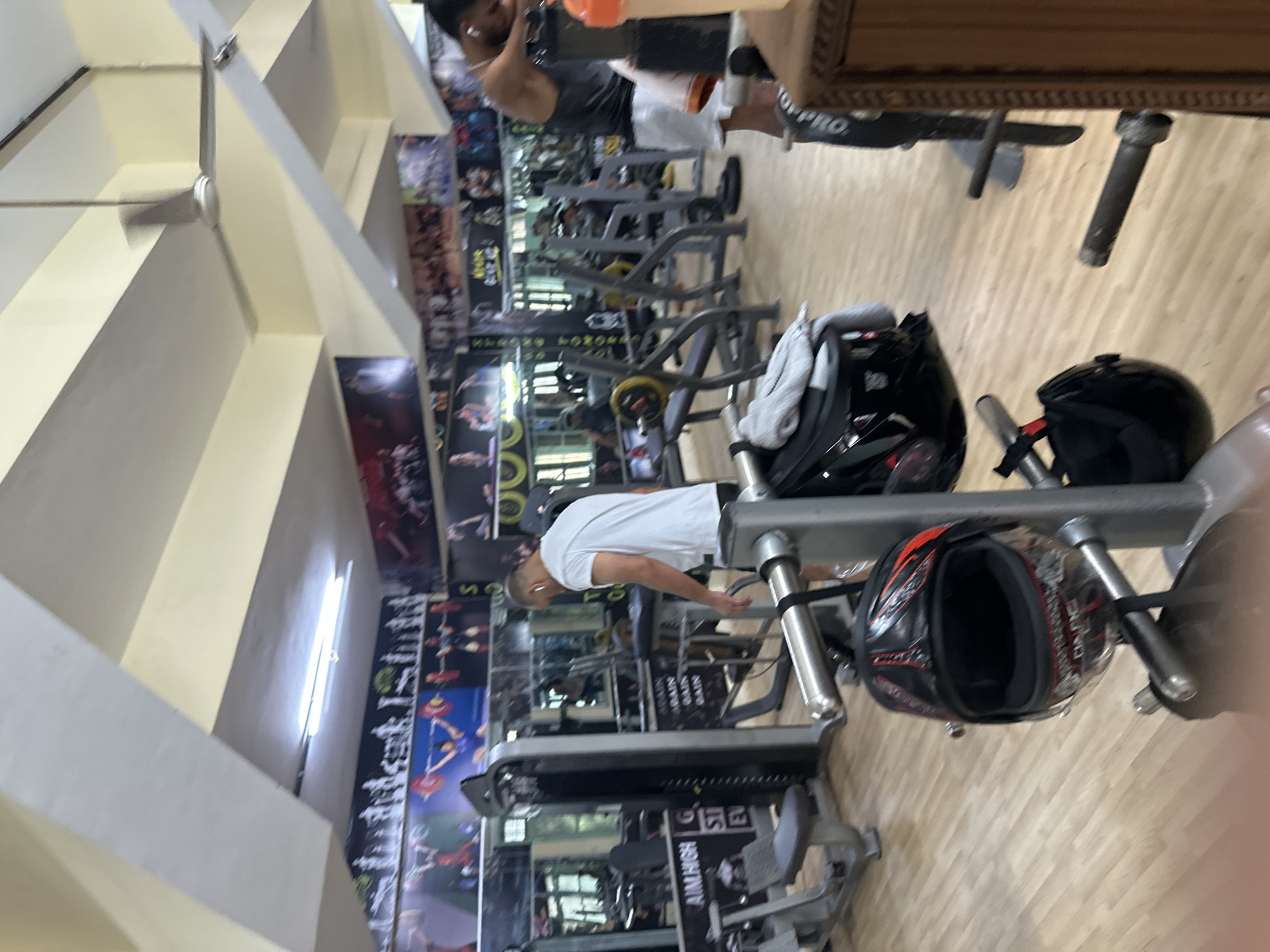 The image depicts the interior of a gym, likely in an urban setting, given the presence of various exercise machines and motivational posters on the walls. The gym appears to be well-equipped, with a variety of weight machines and free weights visible. The flooring is light-colored wood, and the ceiling has a suspended white structure with lights and a ceiling fan.
In the foreground, a black and red motorcycle helmet is hanging on a piece of gym equipment. To the right, a portion of a wooden cabinet is visible. In the middle ground, a man wearing a white t-shirt and earphones is standing and looking down, appearing to be engaged in some activity or resting. Further in the background, another man in a grey tank top and white shorts is leaning on gym equipment, possibly taking a break. Other gym-goers are present but are blurry or out of focus. The overall atmosphere suggests a typical workout session in a commercial gym.