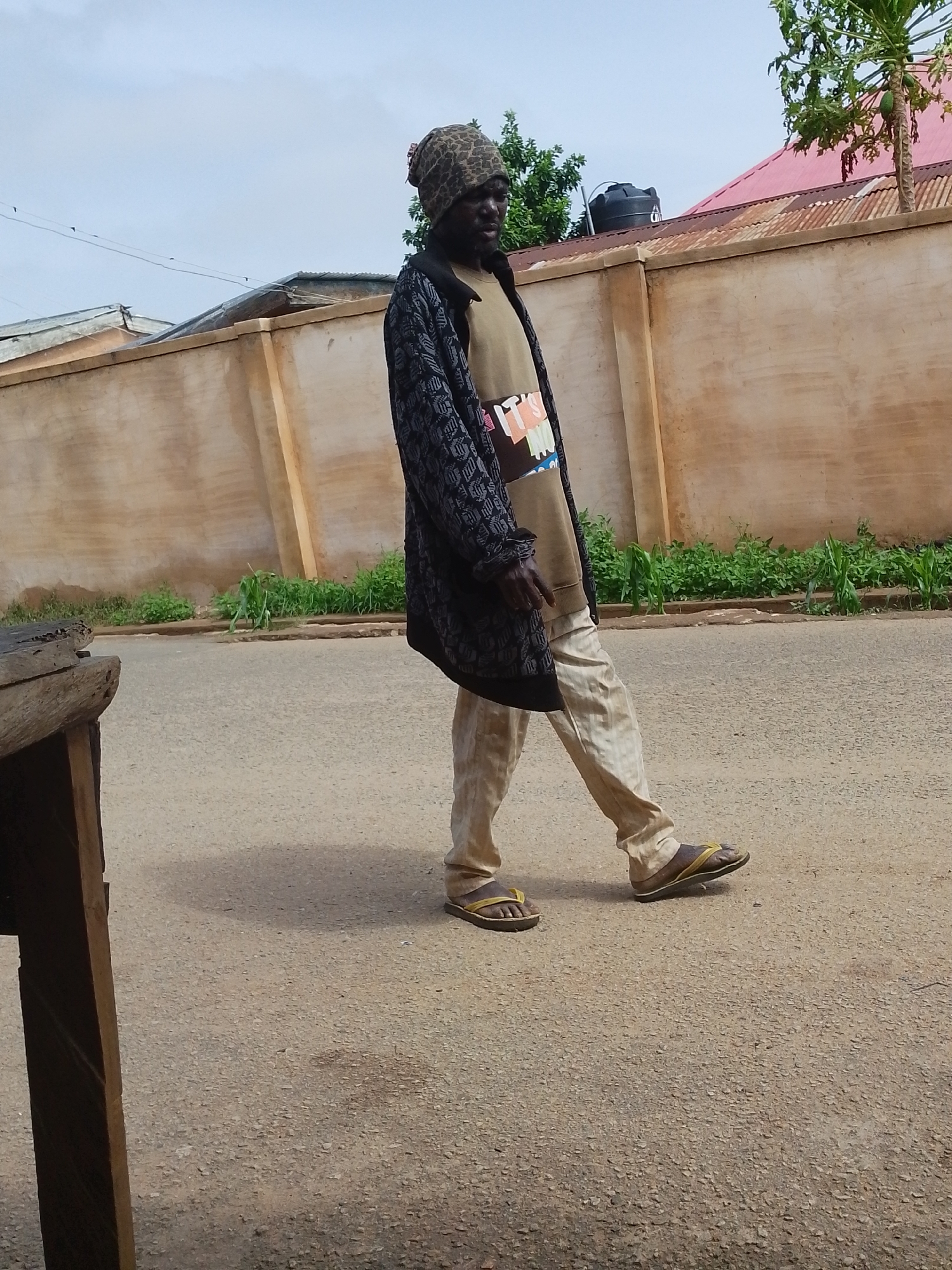 A man is walking on a road in what appears to be a residential area in Jos, Nigeria. The man is wearing a patterned beanie, a long dark coat over a light-colored shirt with a graphic, light-colored pants, and sandals. He is walking from left to right in the frame. To his left is a wooden structure (possibly a table or stall). Behind him is a long concrete wall, and beyond that, houses with corrugated iron roofs. Some greenery is visible along the base of the wall. A tree is partially visible in the upper right corner. The road surface is asphalt and appears to be well-worn.
