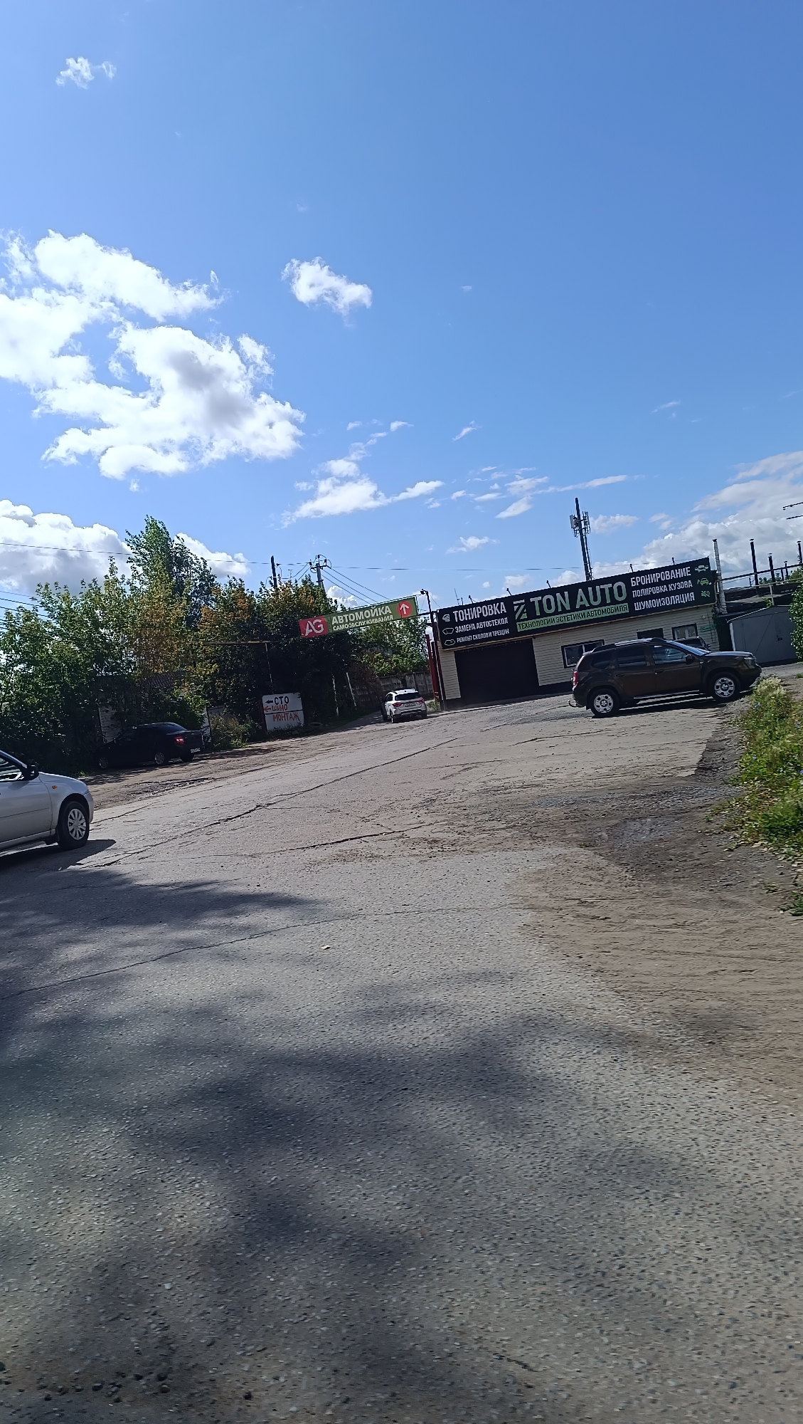 The image shows a street scene with several cars and buildings. In the foreground, a silver sedan is parked on the left side of the road. Further down the road, a black sedan is parked on the right, and a white SUV is driving away from the camera. A brown SUV is parked in front of a building with a large sign that reads "TON AUTO" and lists various automotive services. Another building in the background has a sign that says "АВТОМОЙКА" (car wash). The sky is blue with scattered clouds. The overall setting appears to be an industrial or commercial area. The presence of Russian language signs suggests the location is in Russia, possibly in the city of Chaikovsky, given the prompt. The image is taken during daylight hours under a clear sky.