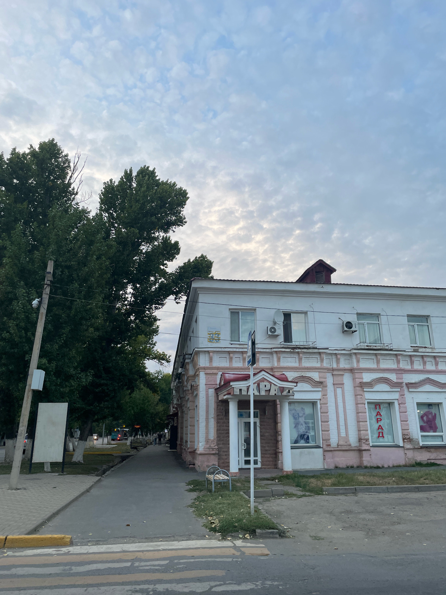 The image shows a street scene in the city of Oral, Kazakhstan, on a cloudy day. A two-story building with a pink and white facade dominates the right side of the frame. The building appears to house a clothing store, as indicated by the mannequins in the windows and the word "ПАРАД" (Parade) visible on a sign and a window display. An entrance with a red awning is visible on the left side of the building's storefront.
To the left of the building, a sidewalk runs alongside a street. Several trees with dense green foliage are present on the left side of the sidewalk, with a utility pole and a blank white sign positioned in front of them. In the distance, traffic can be seen on the street, and a few pedestrians are walking along the sidewalk. A crosswalk is visible in the foreground, with yellow and white stripes. The overall environment suggests a commercial or residential area within the city.