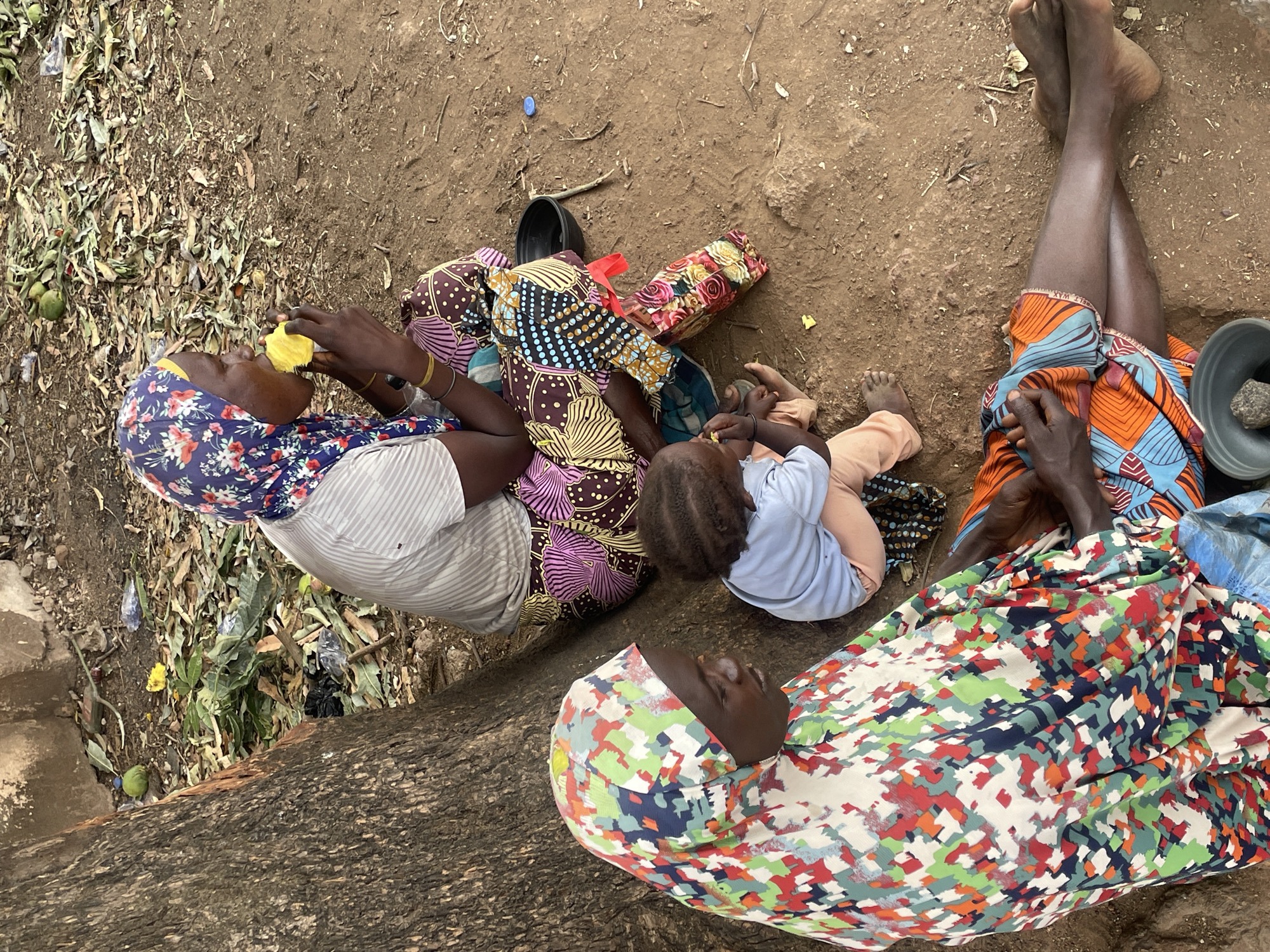 An overhead shot captures four individuals, comprising three adults and one young child, resting on reddish-brown earth, indicative of an outdoor setting in Jos, Nigeria, during daylight hours.
To the left, an adult woman, adorned in a blue floral headscarf and a striped top, is depicted eating a piece of yellow fruit. Adjacent to her, a second adult, wearing a patterned garment with brown, pink, and gold motifs, sits near a young child. The child, dressed in a light blue top and light-colored pants, is also seen consuming a similar yellow fruit. In the right foreground, a third adult is mostly covered by a vibrant, multi-colored geometric patterned fabric, suggesting they are lying or reclining. Further to the right, the bare, crossed legs of another adult are visible, clad in an orange and blue patterned fabric.
The ground is dusty and sparsely covered with dry leaves, some green foliage, and visible litter including plastic bags and a blue bottle cap. Other objects scattered nearby include a floral patterned bag, a single black sandal, and a black plastic bowl containing a stone. The overall scene conveys a moment of rest or casual gathering in a natural, unpaved environment. No discernable text is visible.