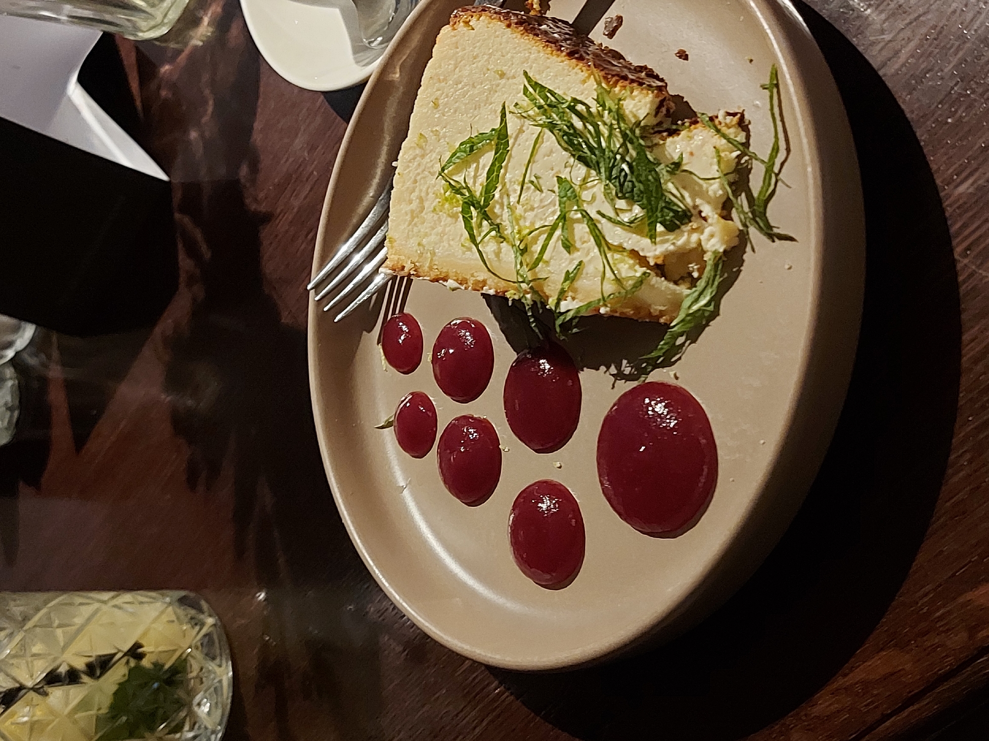 A dessert, likely cheesecake, is presented on a beige plate. It is garnished with strands of green herbs and accompanied by a line of red berry coulis dots. A fork rests on the plate next to the dessert. A glass with a light-colored liquid and green herbs is visible in the bottom left corner of the image. A white saucer is partially visible near the top left corner. The plate is set on a dark wooden surface. The image appears to be taken indoors, possibly in a restaurant. The exact location in Saint Petersburg, Russia, is not discernible from the image.