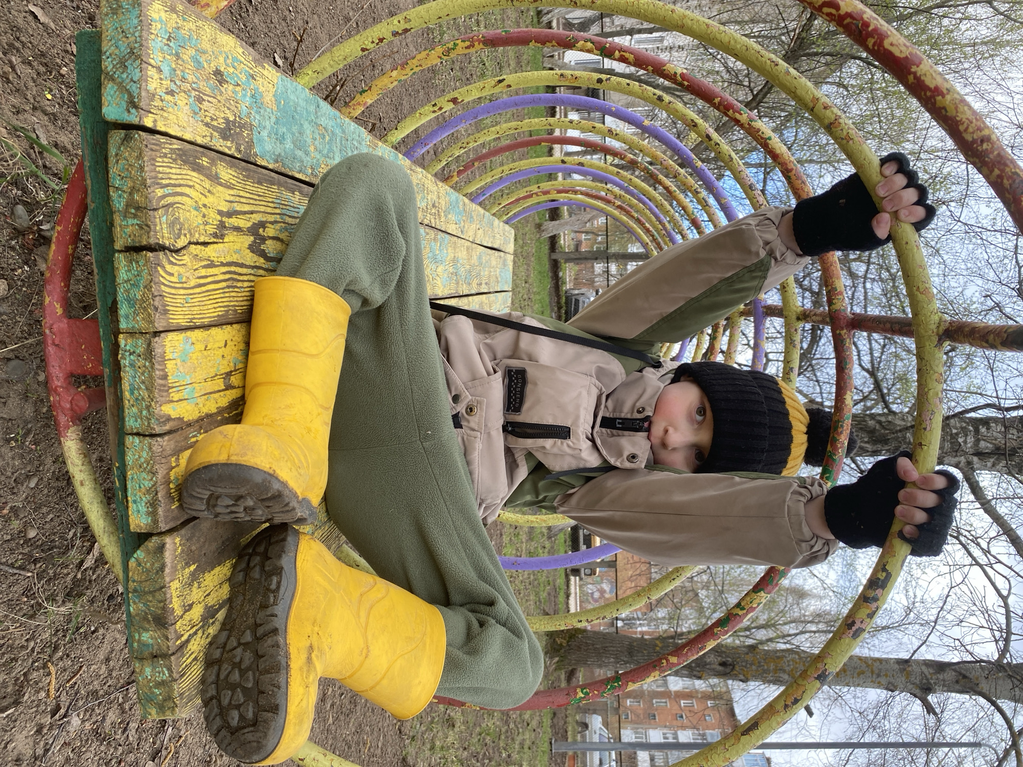 The image, taken in Noviy, Russia, captures a young child positioned horizontally across the frame, lying on a brightly painted yet worn wooden bench within a playground structure. The child is dressed for cool weather, wearing vibrant yellow rubber boots, olive green fleece pants, a beige and olive green hooded jacket, black fingerless gloves, and a black and yellow striped knit hat with a pompom. They gaze directly at the camera with a neutral expression, their arms outstretched to grip the yellow bars of the colorful archway above them.
The playground equipment features a series of arches, or a tunnel, painted in yellow, red, and purple, all showing visible signs of rust and chipped paint, indicating age and use. The wooden bench the child is on has a distressed look, with yellow and blue-green paint peeling to reveal the natural wood grain underneath. In the background, through the bare branches of trees, distant residential buildings are visible under a bright, likely overcast, sky. The ground is dirt and sparse grass, suggesting an outdoor setting. The bare trees imply late winter or early spring. Faint text, possibly "CONSIENO." or similar, is discernible on the child's jacket.
